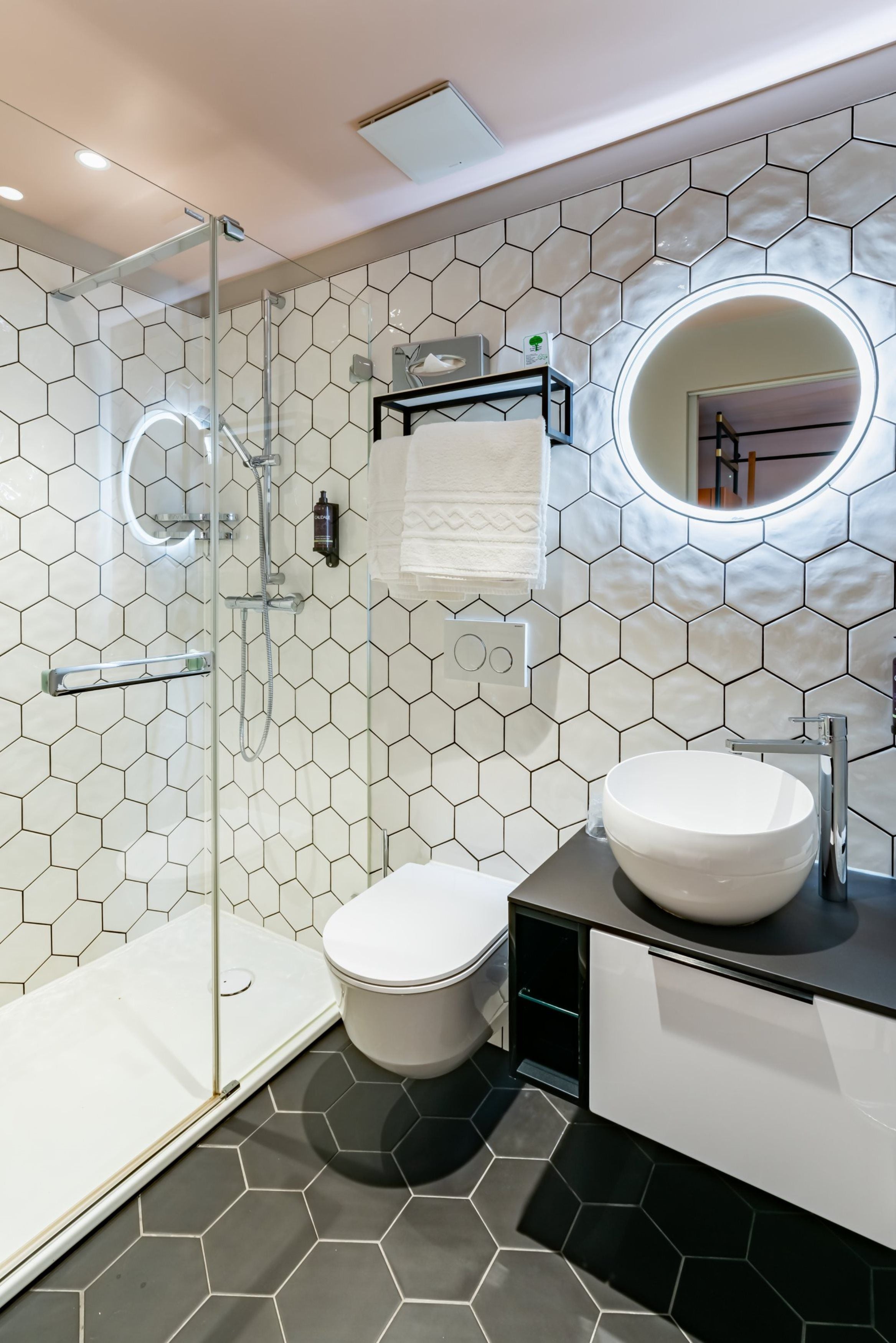 Een moderne badkamer met een betegeling in een honingraatpatroon, een glazen douchecabine, een wit toilet, een wastafel met een ronde spiegel en een handdoekhouder.