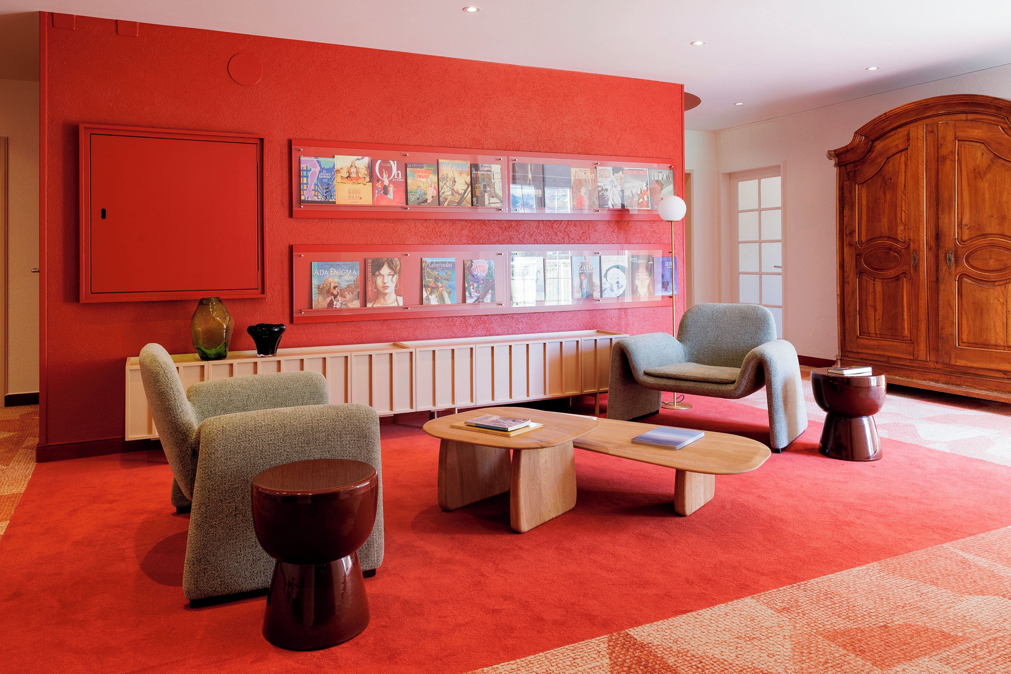 Ein Zimmer mit roten Wänden, einem Holzschrank und einem Bücherregal mit Büchern. Es gibt zwei Sessel und einen Couchtisch.