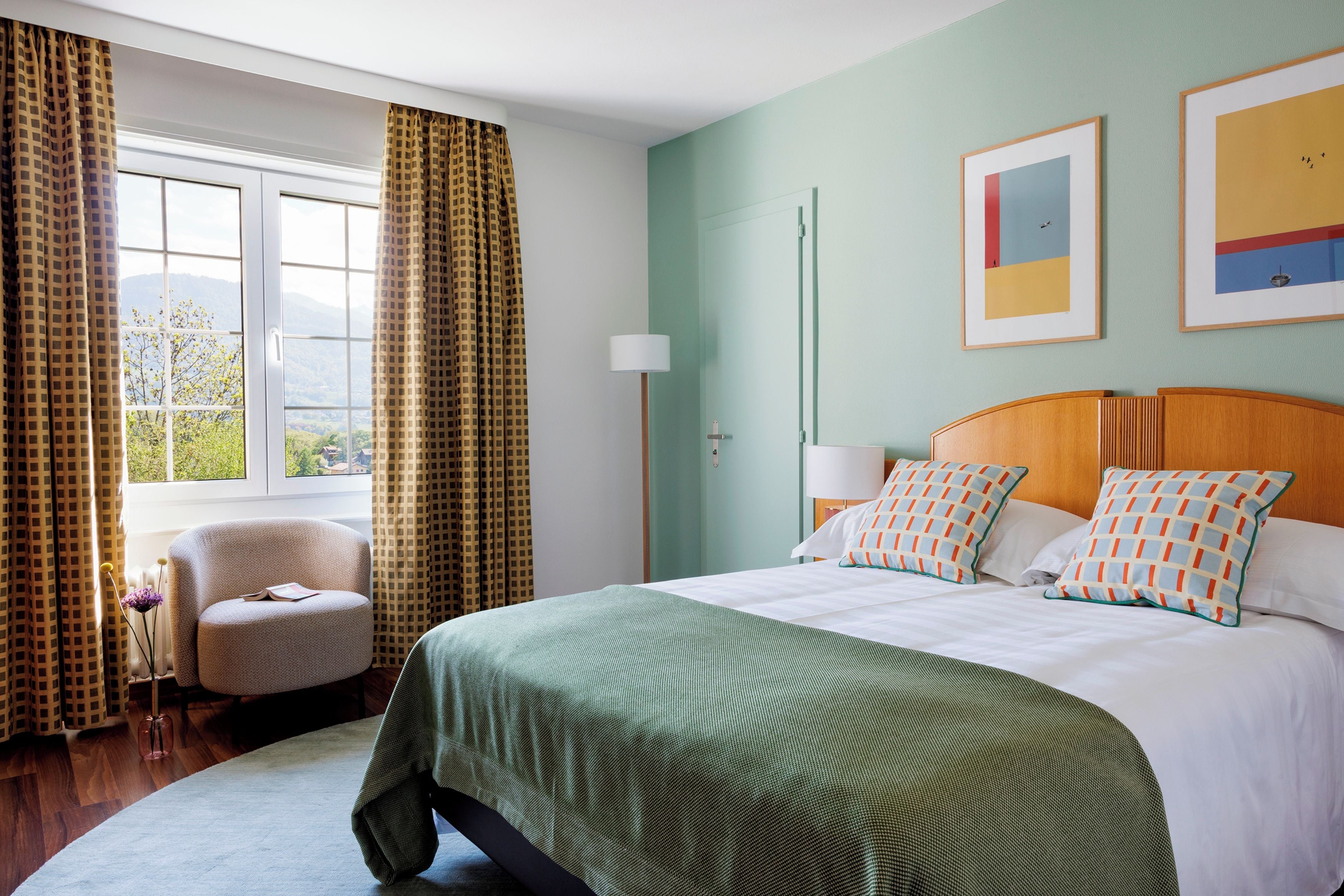 Une chambre d’hôtel avec un grand lit, une chaise, une fenêtre avec des rideaux et des œuvres d’art accrochées au mur.