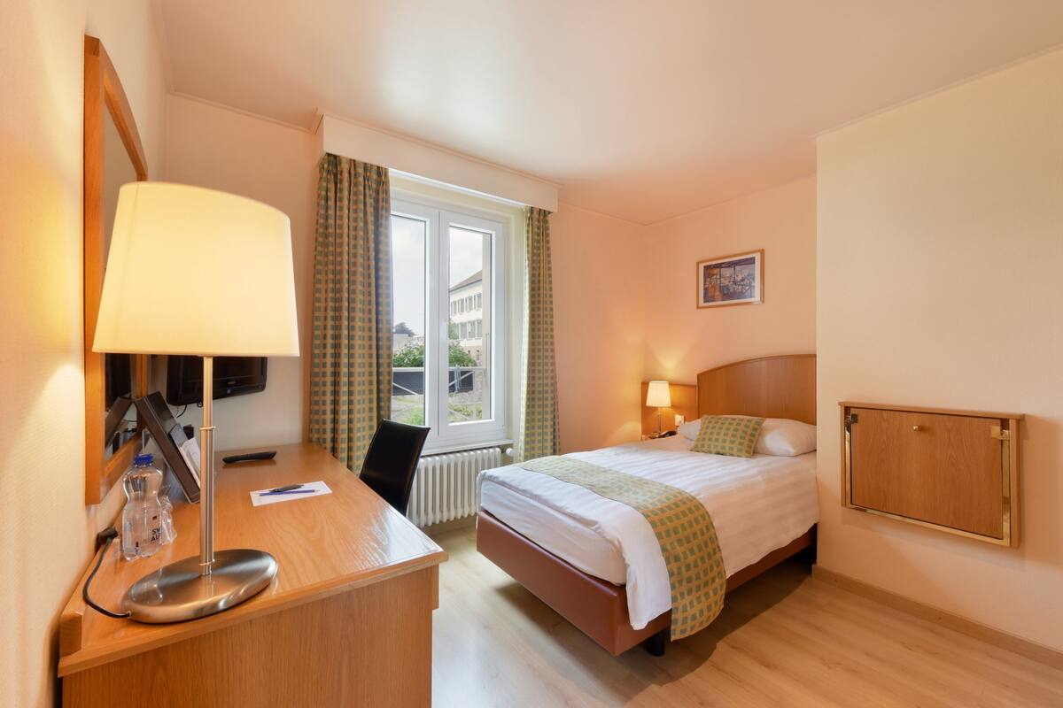 A hotel room with a bed, bedside lamps, a desk with a lamp, a chair, a window with curtains, and a picture on the wall.