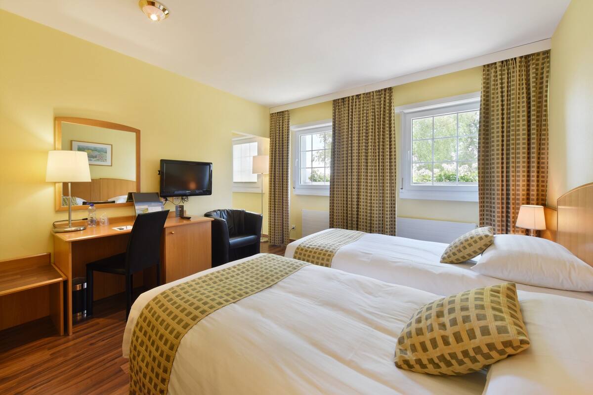 A hotel room with two beds, a desk with a chair, a television, and a window with curtains.