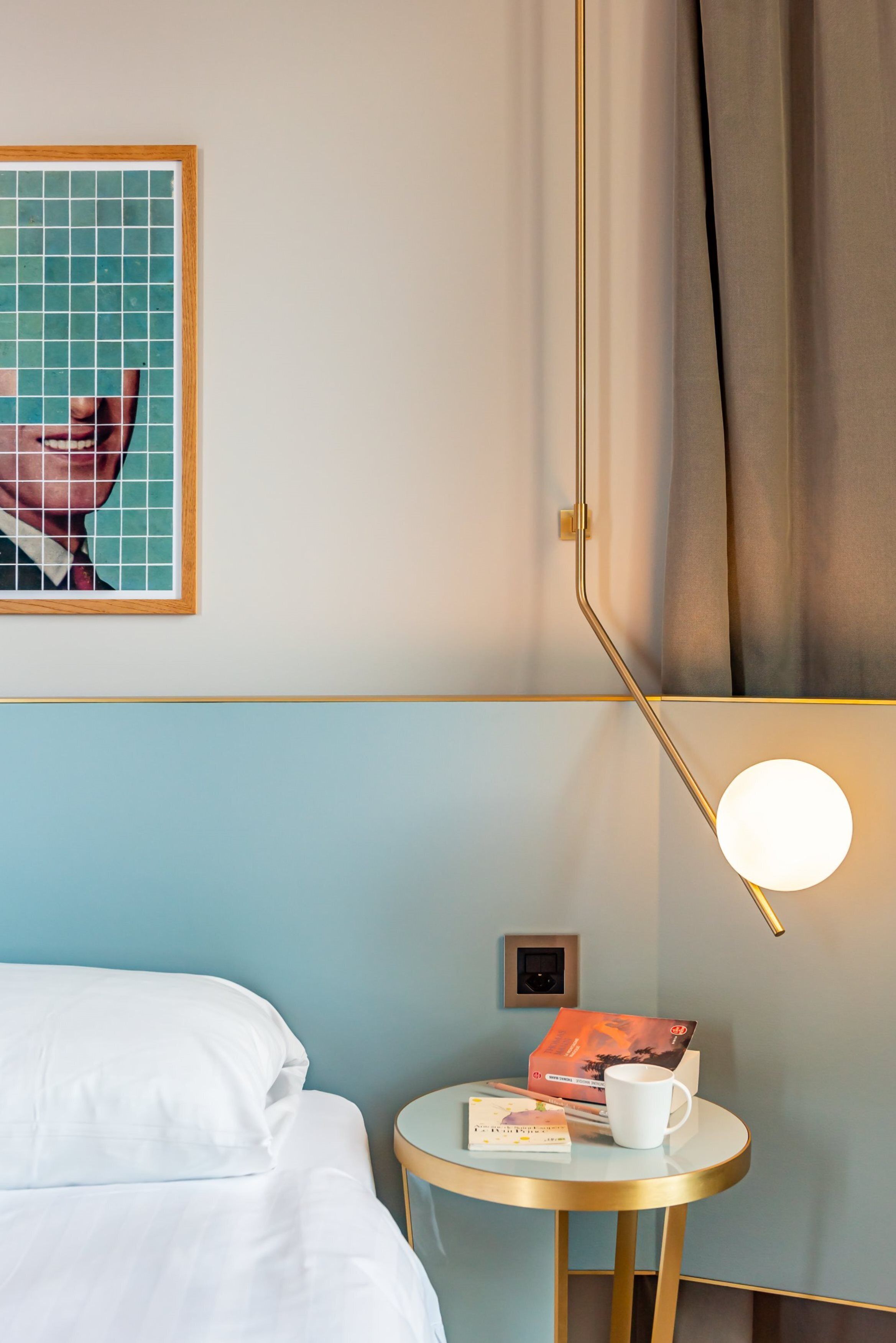 Ein Hotelzimmer mit einem Bett, einem kleinen runden Tisch mit Buch und Tasse, einem gerahmten Bild an der Wand und einer Lampe.