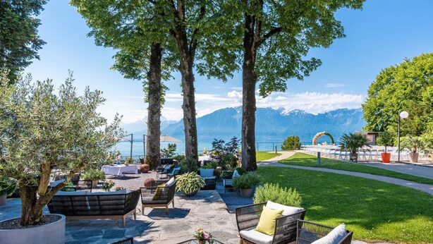An outdoor seating area with wicker furniture, potted plants, and a view of a lake and mountains. - Hotel du Leman (Jongny)