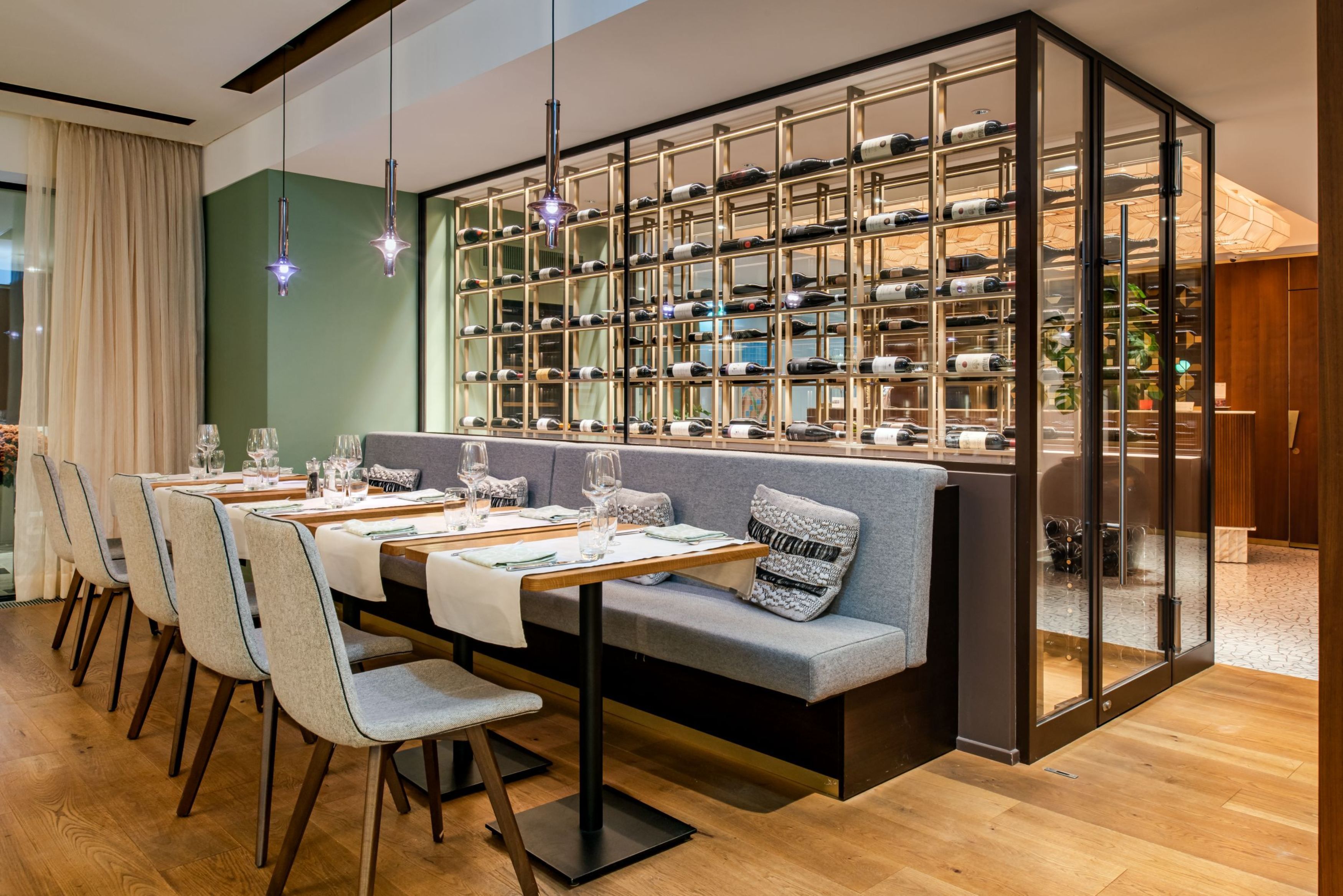 A dining area with a long table, chairs, and a seating area with a sofa. There is a wine rack with bottles behind the sofa.