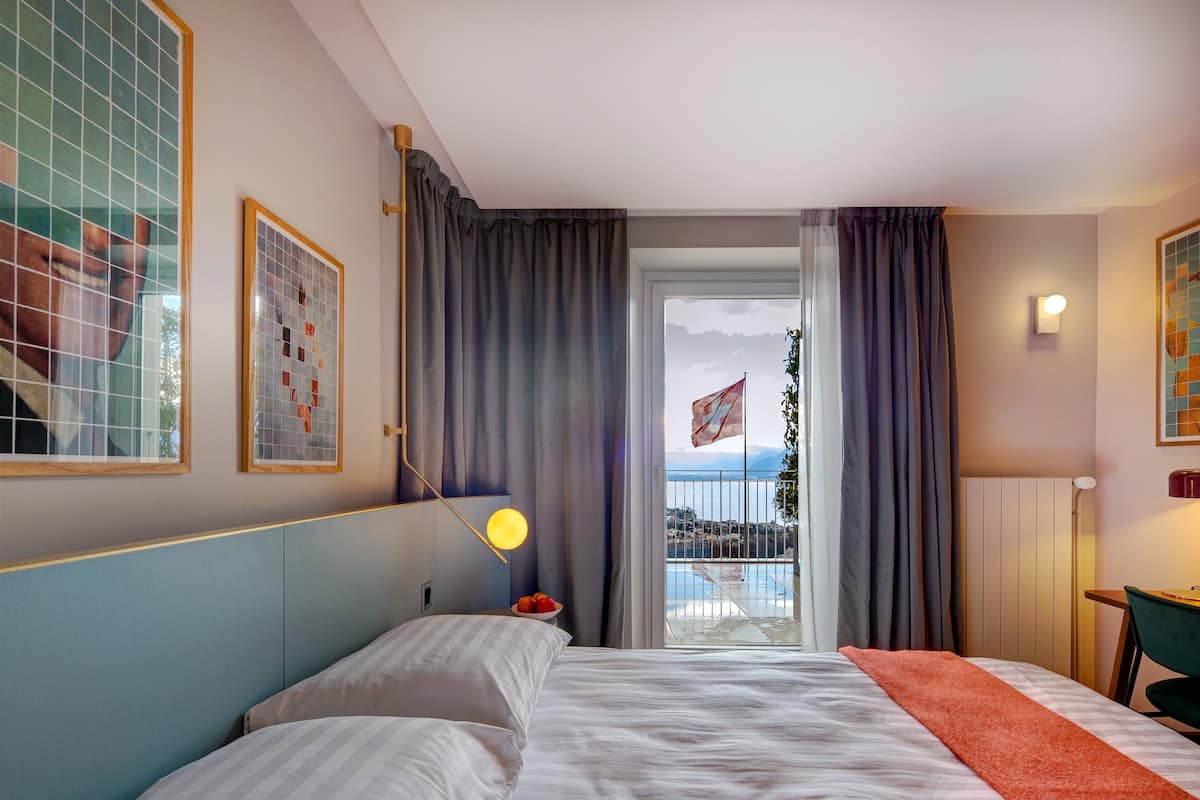 A hotel room with a bed, a nightstand, a lamp, a window with curtains, and a view of a balcony with a flag.