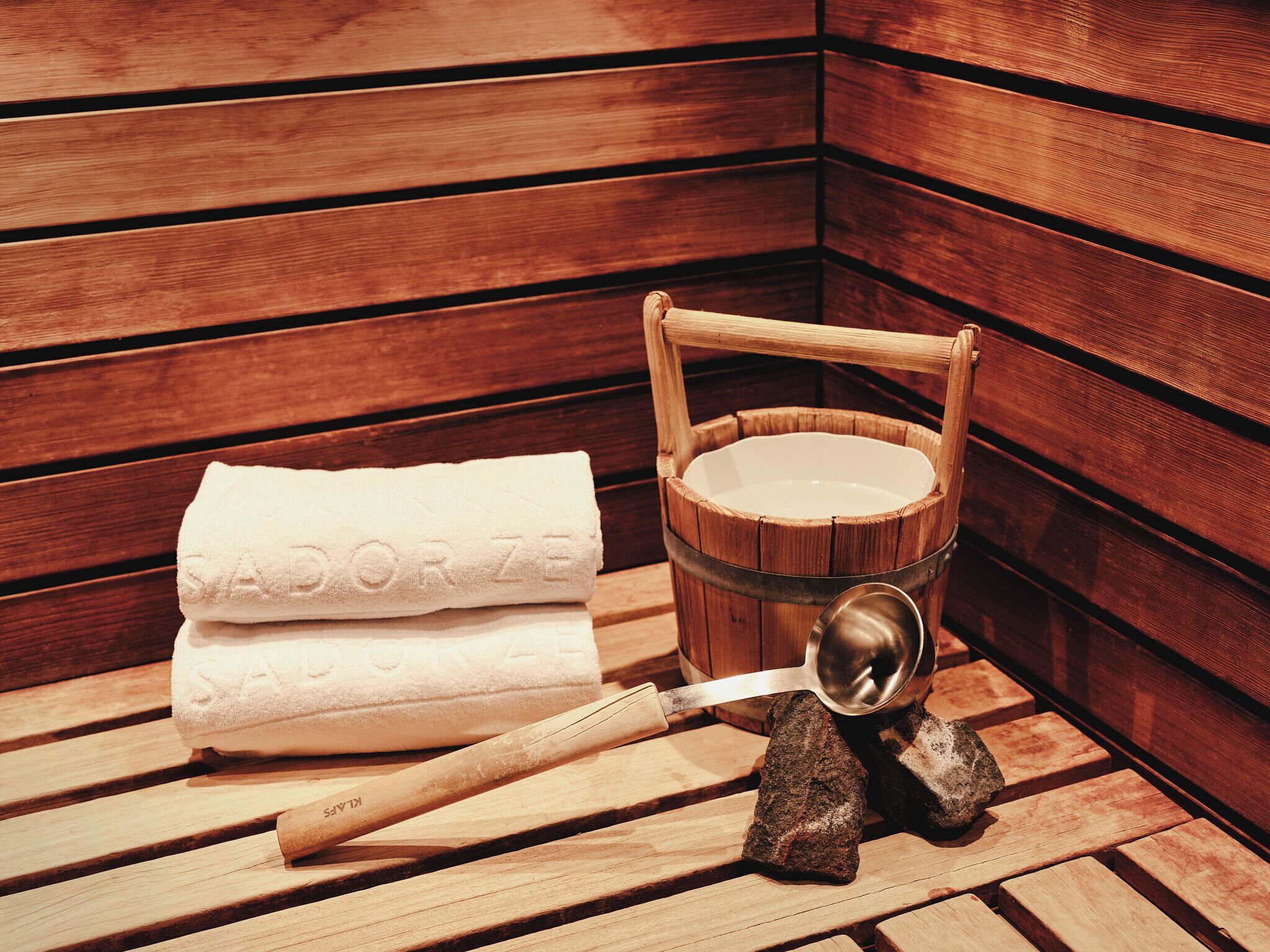 A wooden sauna with a wooden bench, a towel, a wooden bucket, and a ladle.