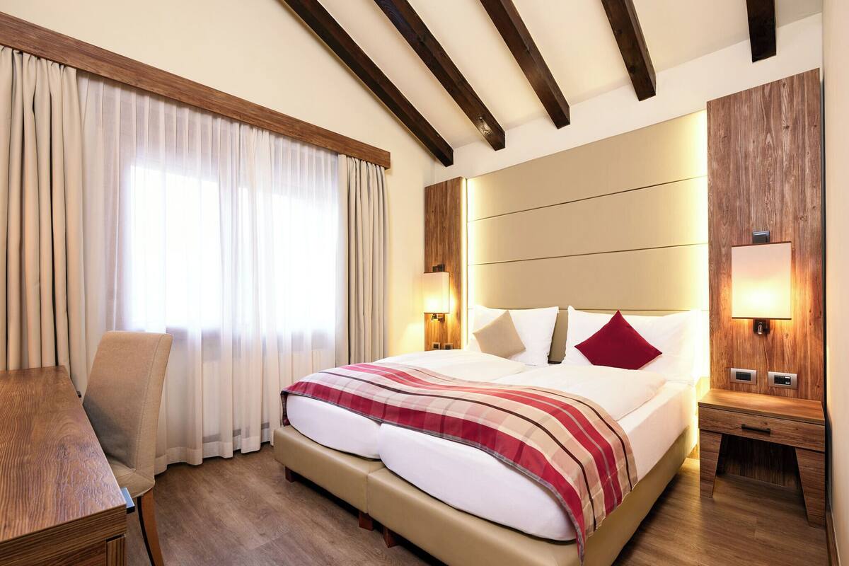 A hotel room with a large bed, wooden headboard, bedside tables with lamps, a wooden desk, and a chair.