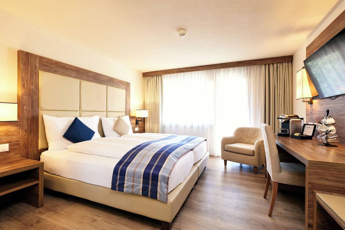 A hotel room with a large bed, a desk with a coffee machine, and a TV mounted on the wall.