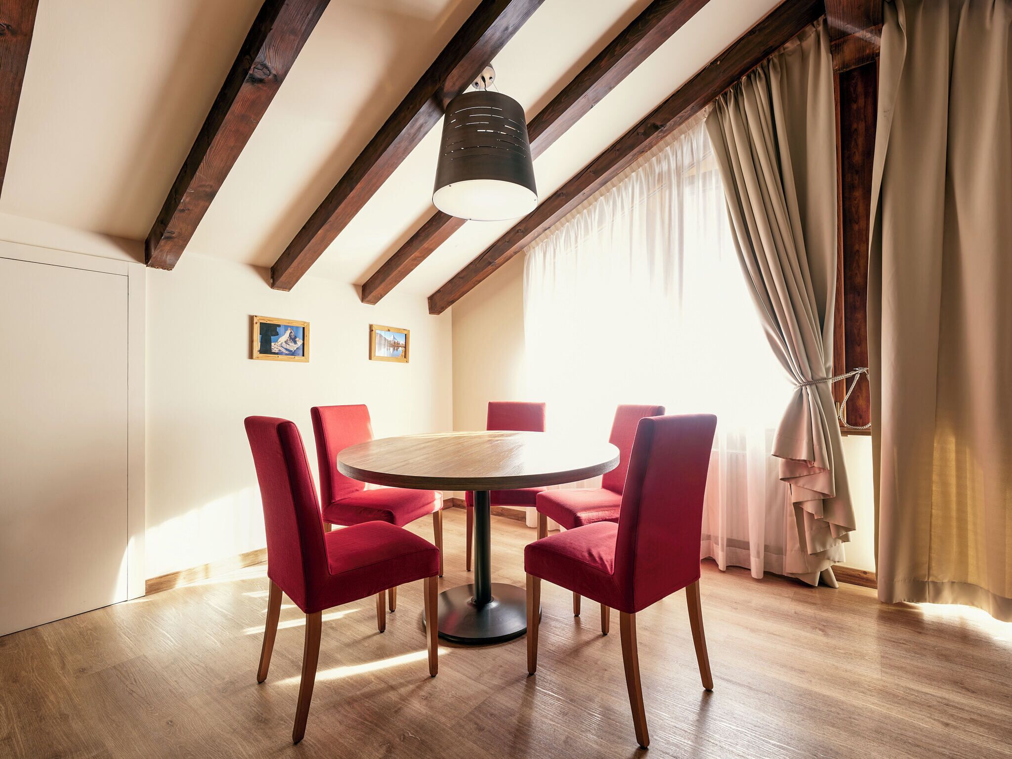 Een eethoek met een ronde tafel, rode stoelen, houten balken en een hanglamp.