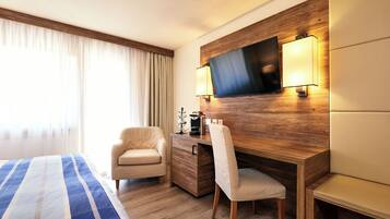 Una camera d'hotel con un letto, una scrivania, una sedia, una TV fissata a una parete in legno e una finestra con tende.