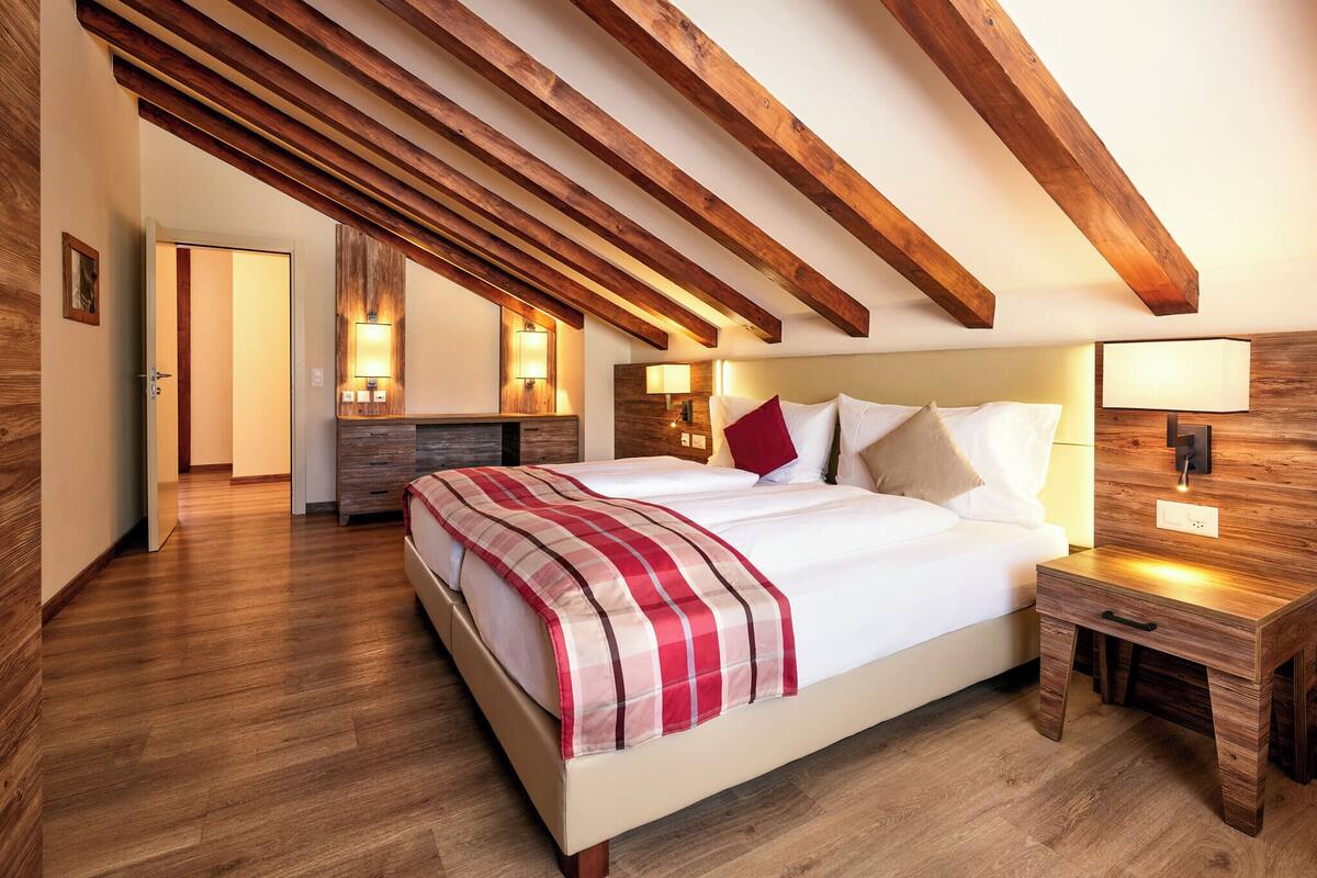 A bedroom with a wooden ceiling, a large bed with a plaid blanket, bedside tables, and wall-mounted lamps.