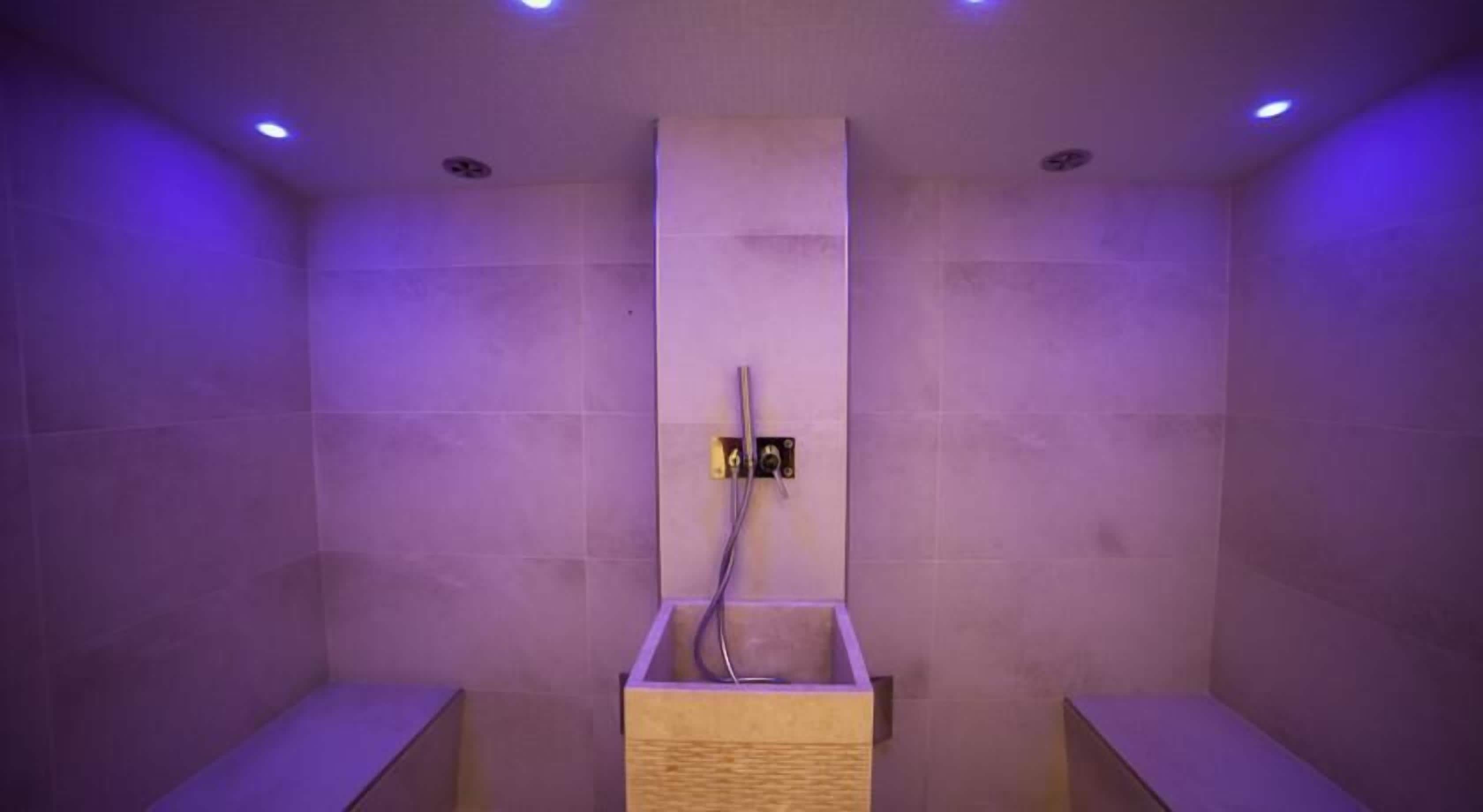 A modern shower area with a wooden bench, a wall-mounted control panel, and recessed ceiling lighting.