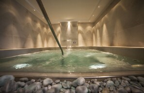A spa bath with a waterfall feature, surrounded by stone walls and a ceiling light.
