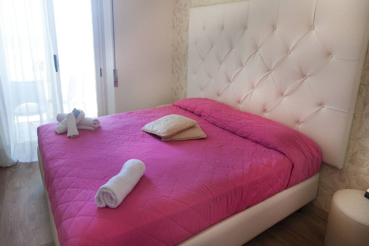 A bed with a tufted headboard, a pink quilted bedspread, and rolled towels on the side.