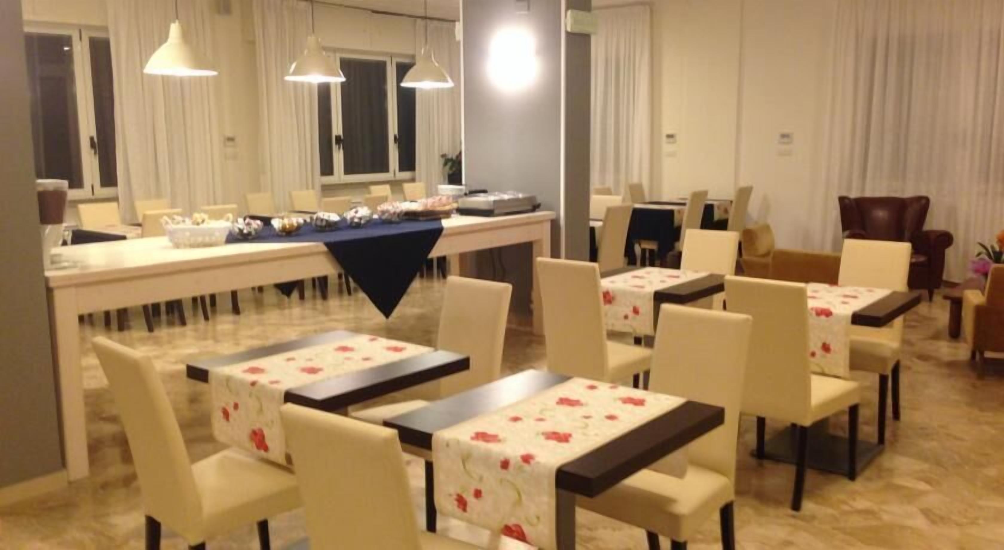 A hotel dining area with multiple tables and chairs, a buffet setup, and large windows with curtains.