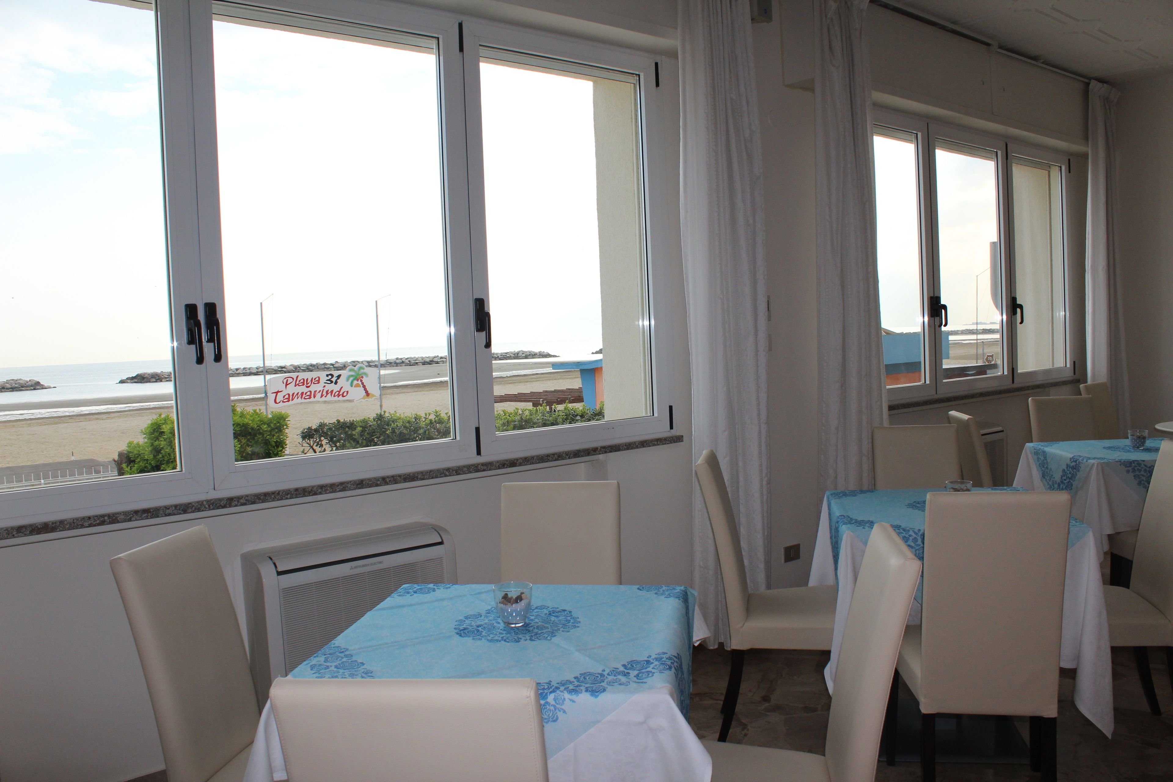 A dining area with tables and chairs, large windows with curtains, and a view of the beach.