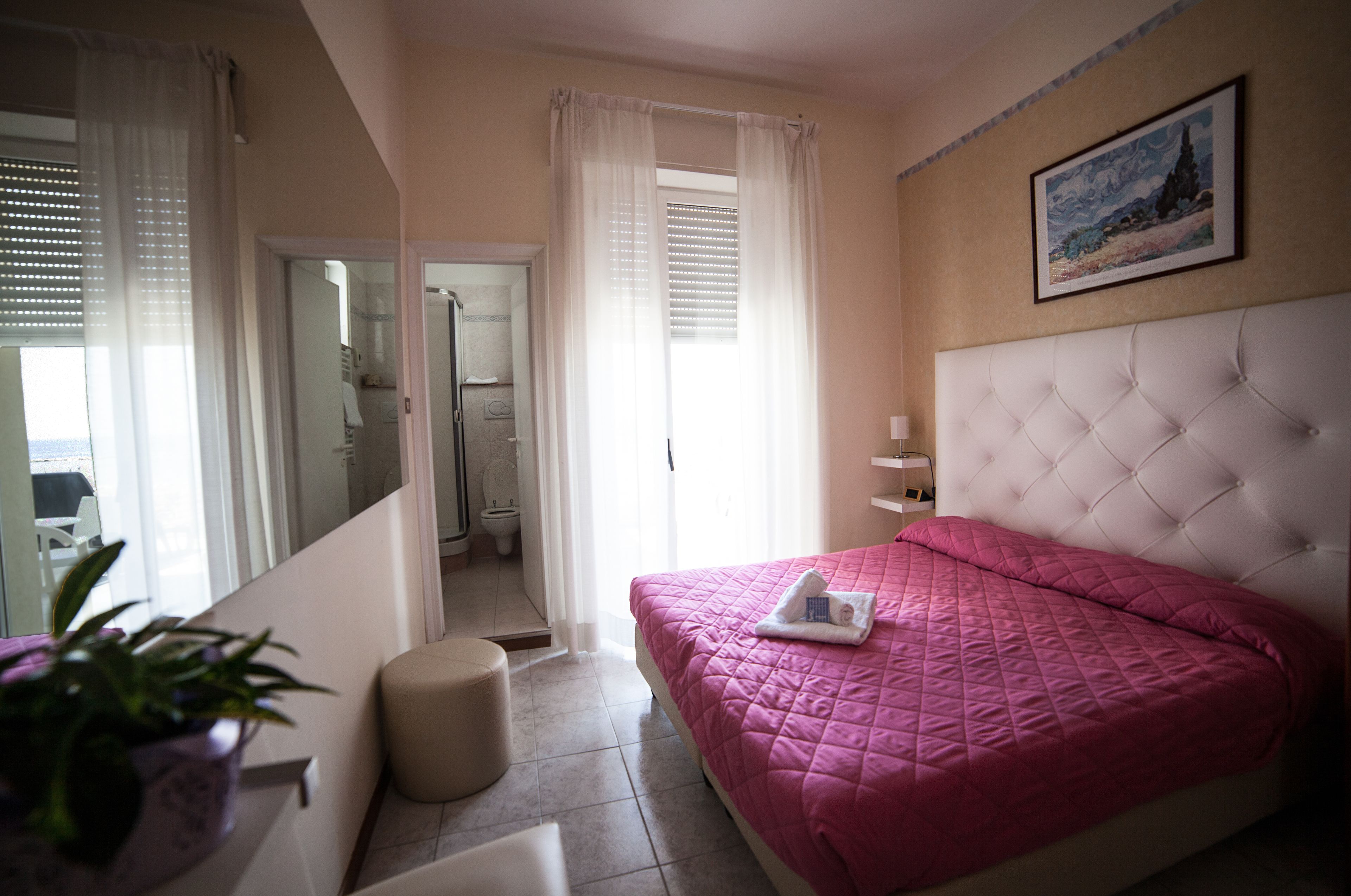 Ein Schlafzimmer mit einer rosa gesteppten Bettdecke, einem weißen, geknöpften Kopfteil, einem Gemälde an der Wand und einem Blumentopf am Fenster.