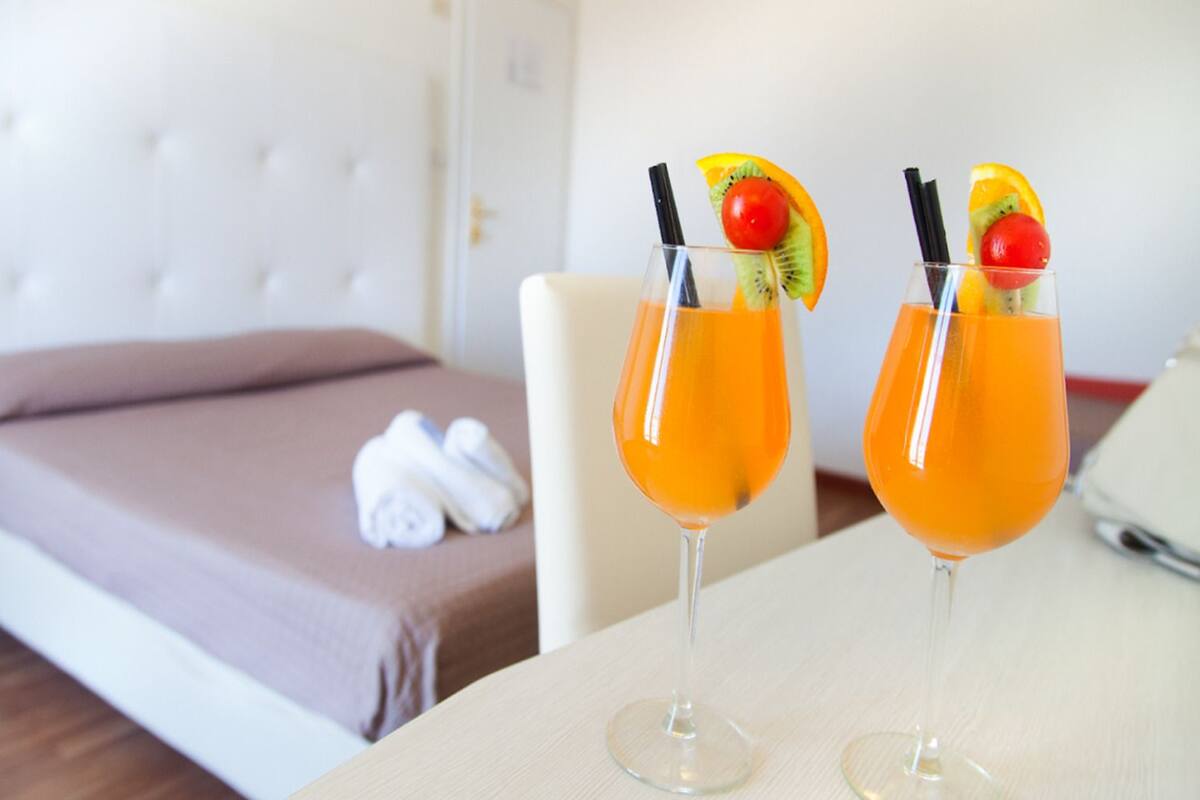 Two cocktails with fruit garnishes on a table in a hotel room with a bed and towels in the background.