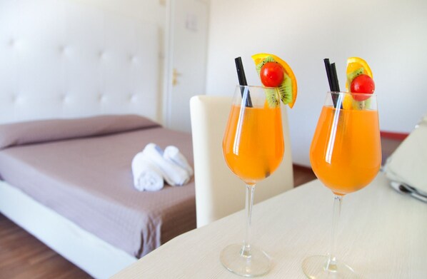 Two cocktails with fruit garnishes on a table in a hotel room with a bed and towels in the background.