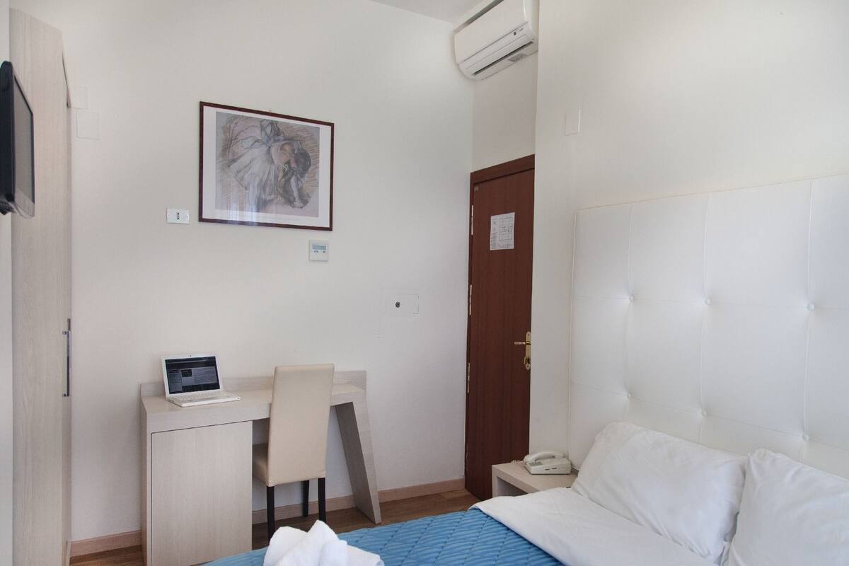 A hotel room with a bed, a desk with a laptop, a chair, a bedside table with a phone, and a door.