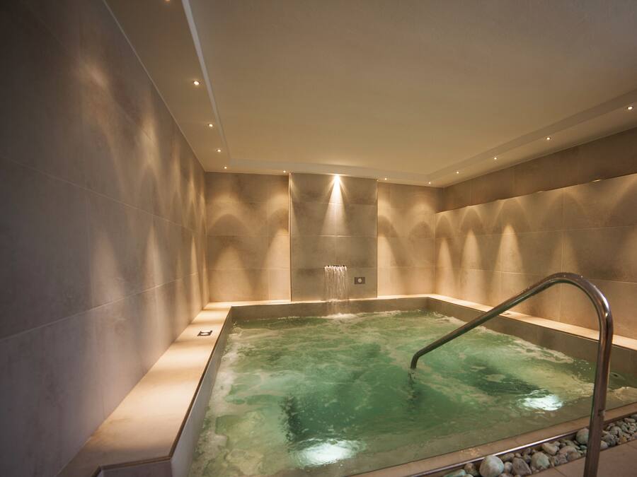 A modern spa room with a rectangular hot tub, a metal ladder, and a wall-mounted water feature.