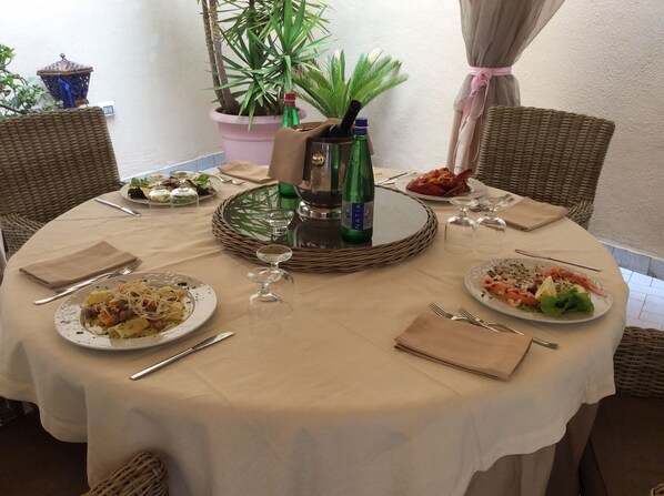A round dining table with a beige tablecloth, surrounded by wicker chairs. There are plates of food, a bottle of NATA, and a decorative lantern. - Hotel Castello dei Principi (Capaccio-Paestum)