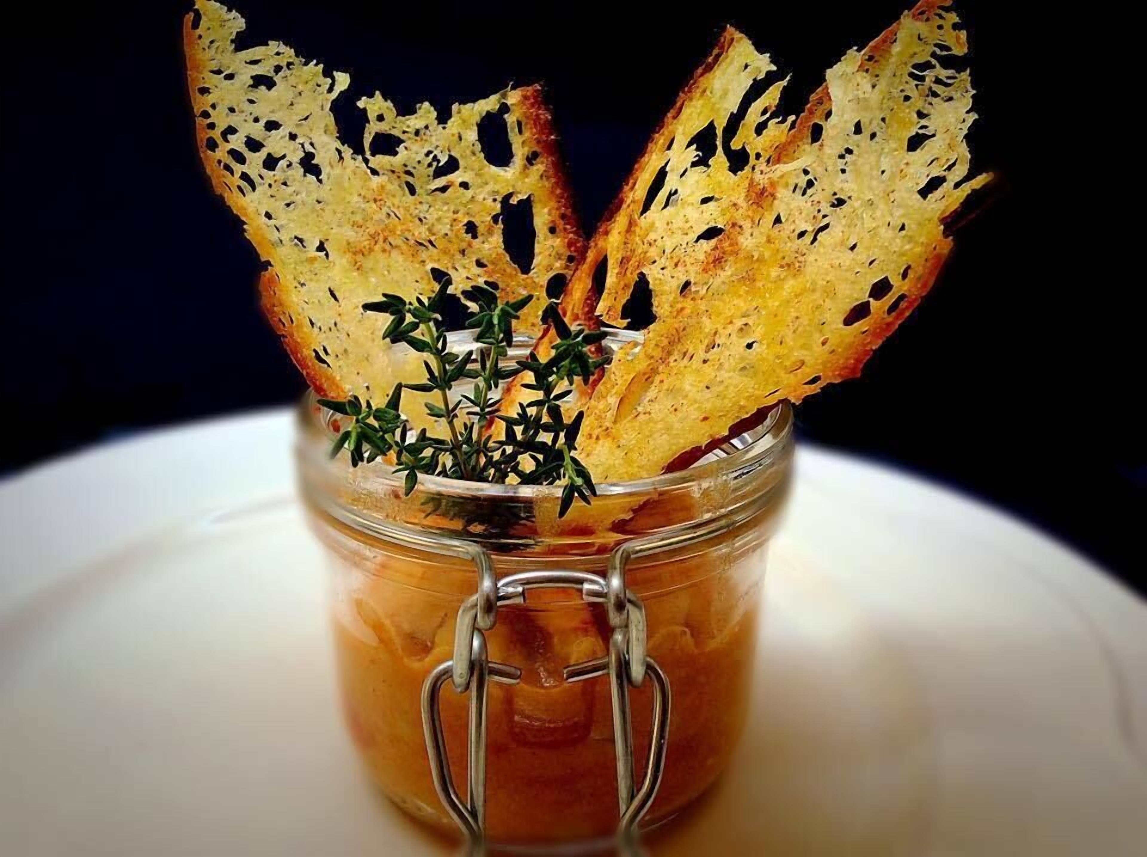 Ein Glasgefäß mit Metallverschluss, das eine dunkle Flüssigkeit, vermutlich eine Soße oder Marinade, enthält, mit gerösteten Brocken und frischen Kräutern belegt.
