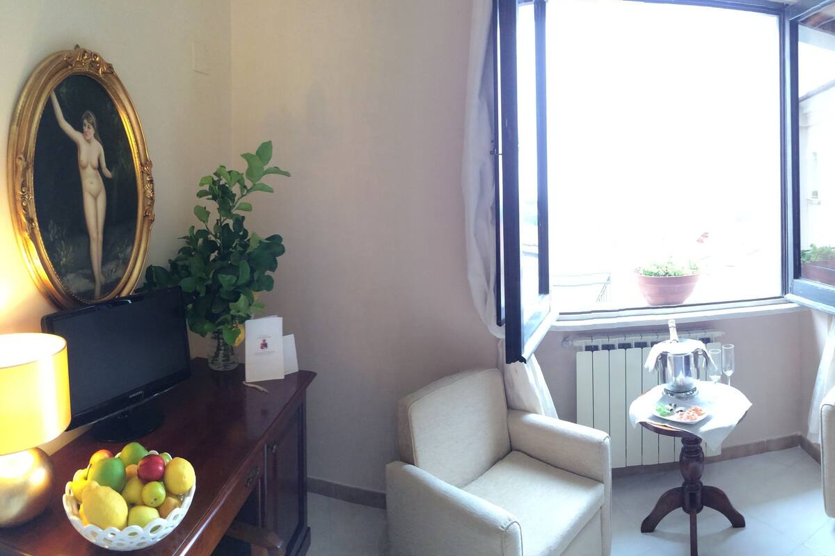Un salon avec deux fauteuils blancs, une petite table sur laquelle se trouvent une théière et des verres, une plante en pot, un tableau encadré au mur et un téléviseur posé sur un meuble en bois.