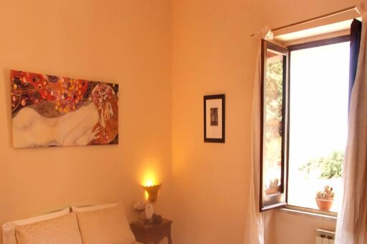 Une chambre à coucher avec un lit, une table de chevet, une lampe, une fenêtre avec des rideaux et un tableau accroché au mur.