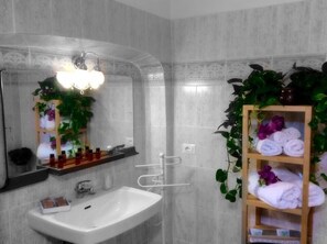 A bathroom with a white sink, a mirror, and a shelf with towels and plants. - Hotel Castello dei Principi (Capaccio-Paestum)