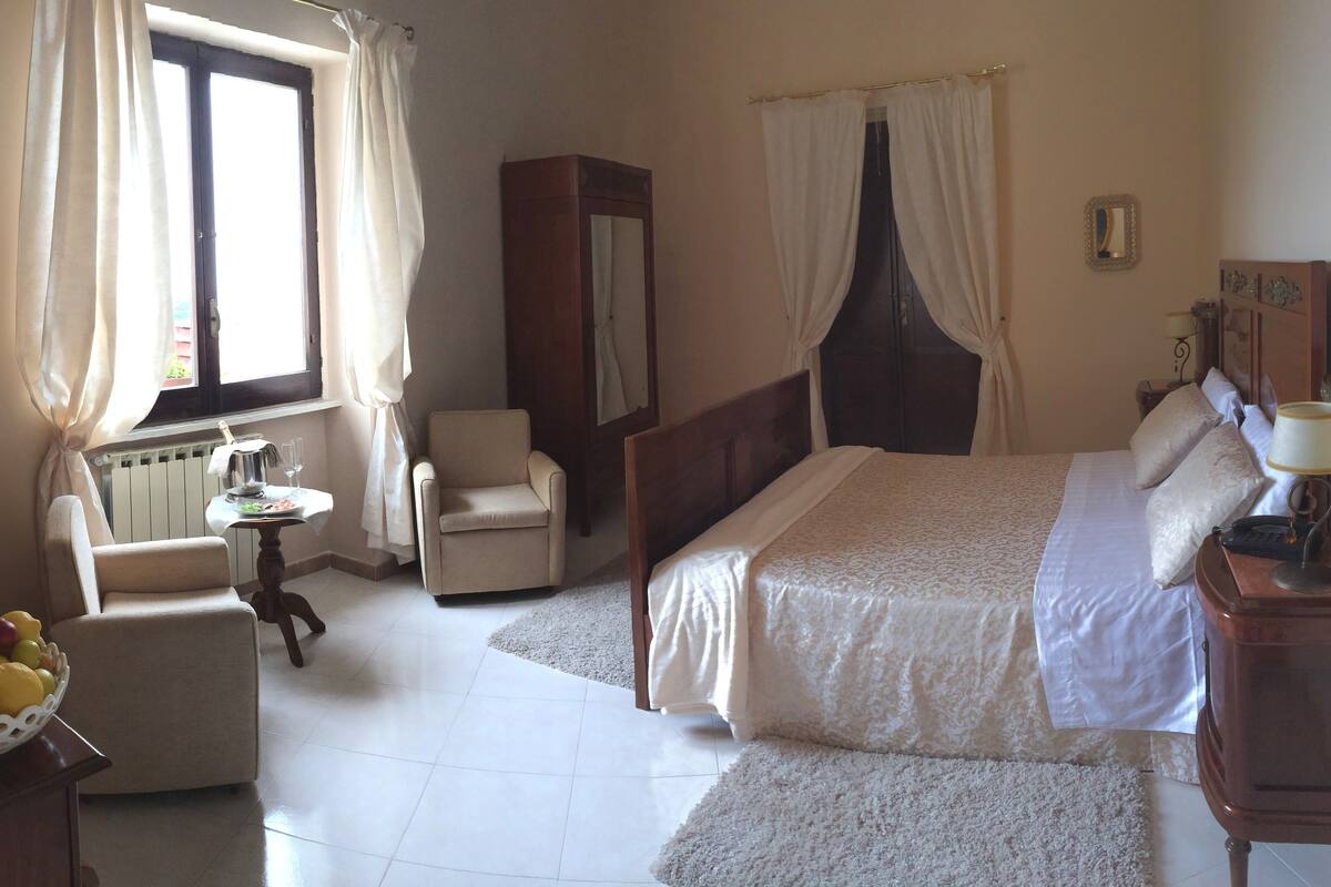 Une chambre d’hôtel avec un lit, une chaise, une petite table avec une lampe et une fenêtre avec des rideaux.