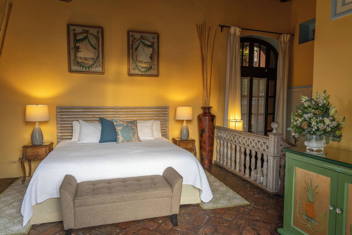 Ein Schlafzimmer mit einem großen Bett, zwei Nachttischlampen, einer Bank, einem hölzernen Kopfteil und einem dekorativen Schrank mit Ananas-Motiv.