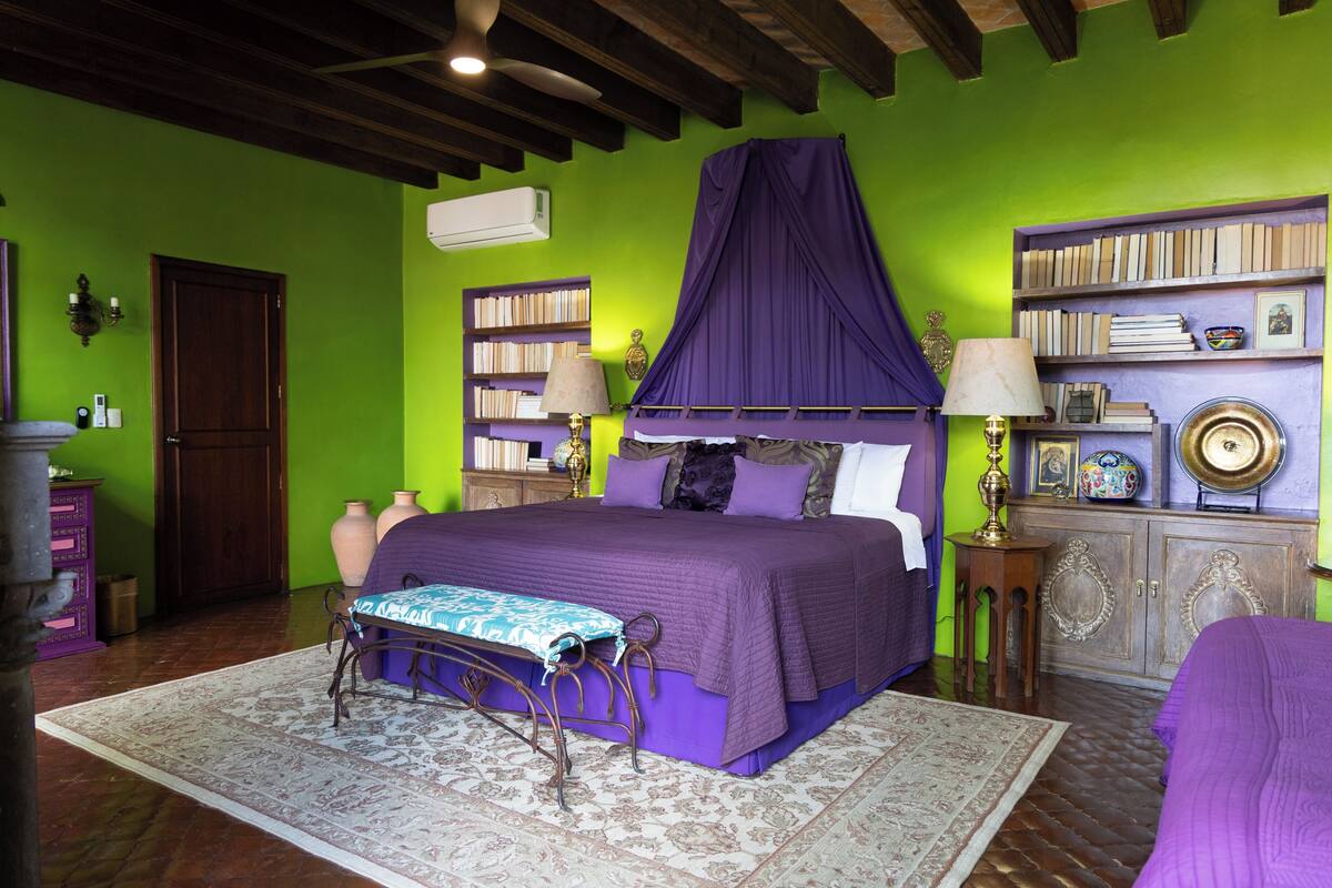 Ein Schlafzimmer mit Bett, Nachttisch, Bücherregal und einem violetten Baldachin.
