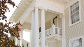 Une maison à deux étages dotée d’un porche soutenu par des colonnes blanches, d’un rez-de-chaussée en briques et d’un balcon au deuxième étage.