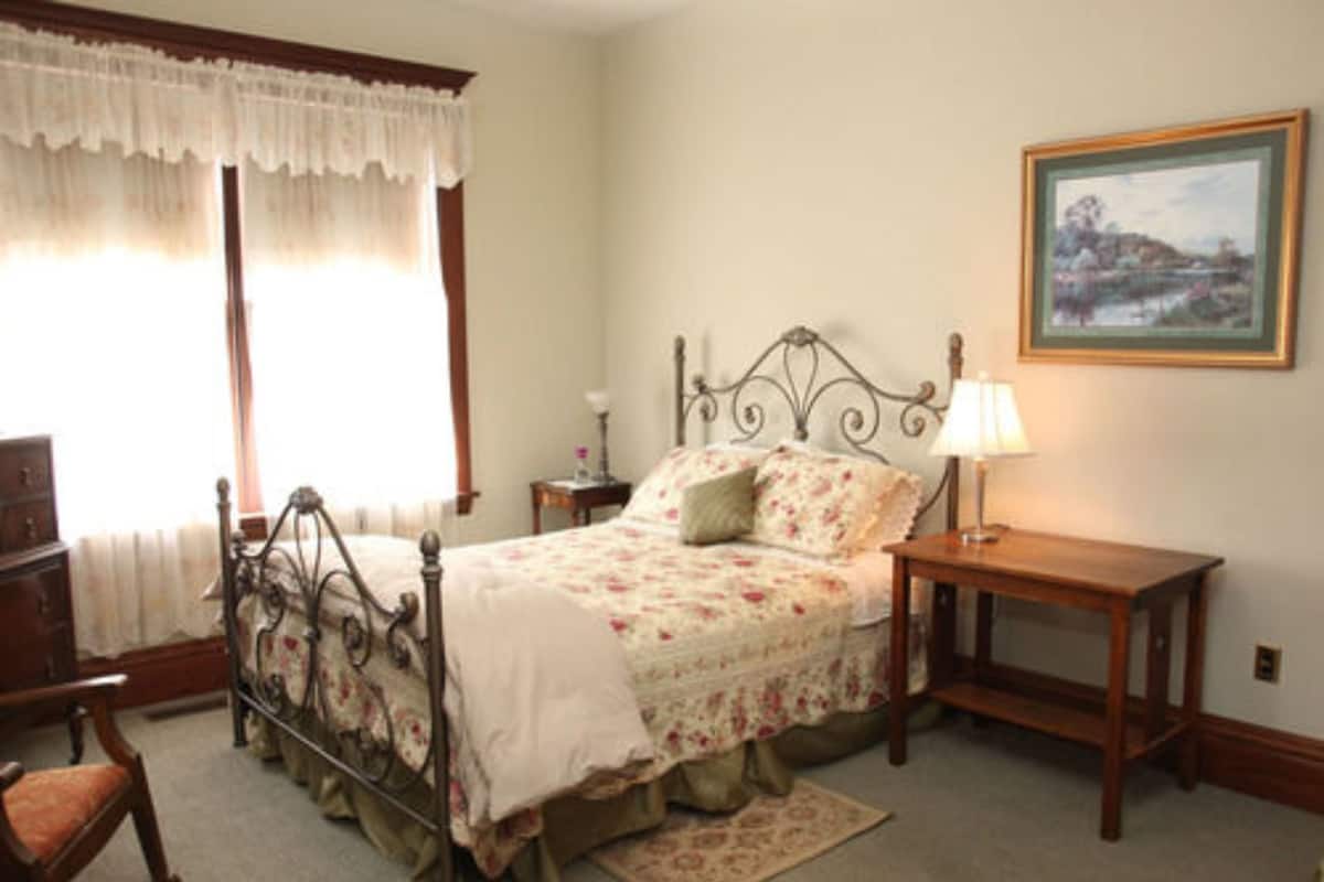 A bedroom with a four-poster bed, a nightstand, a chair, and a painting on the wall.