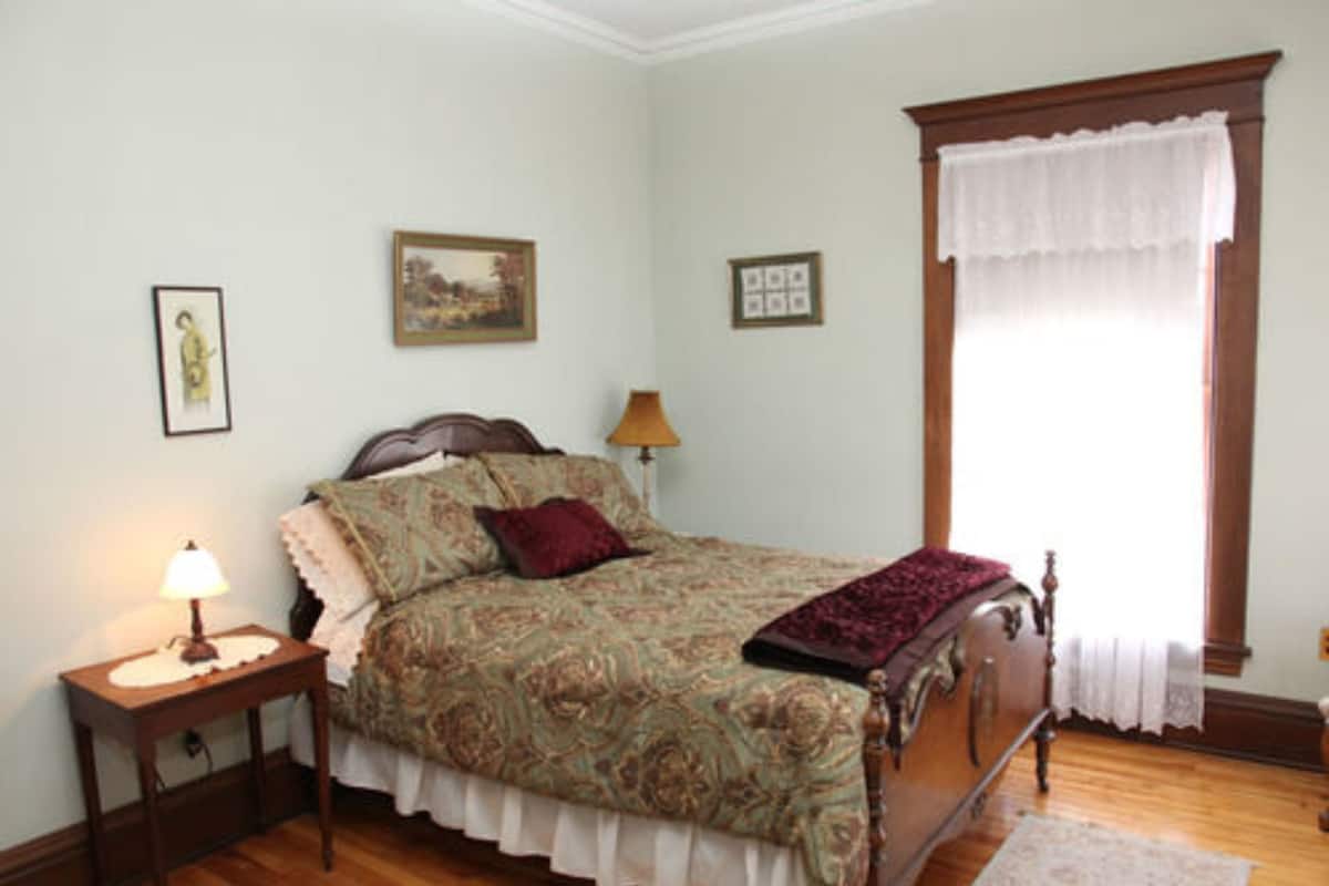 A bedroom with a bed, a nightstand, a lamp, a window with a white curtain, and framed pictures on the wall.