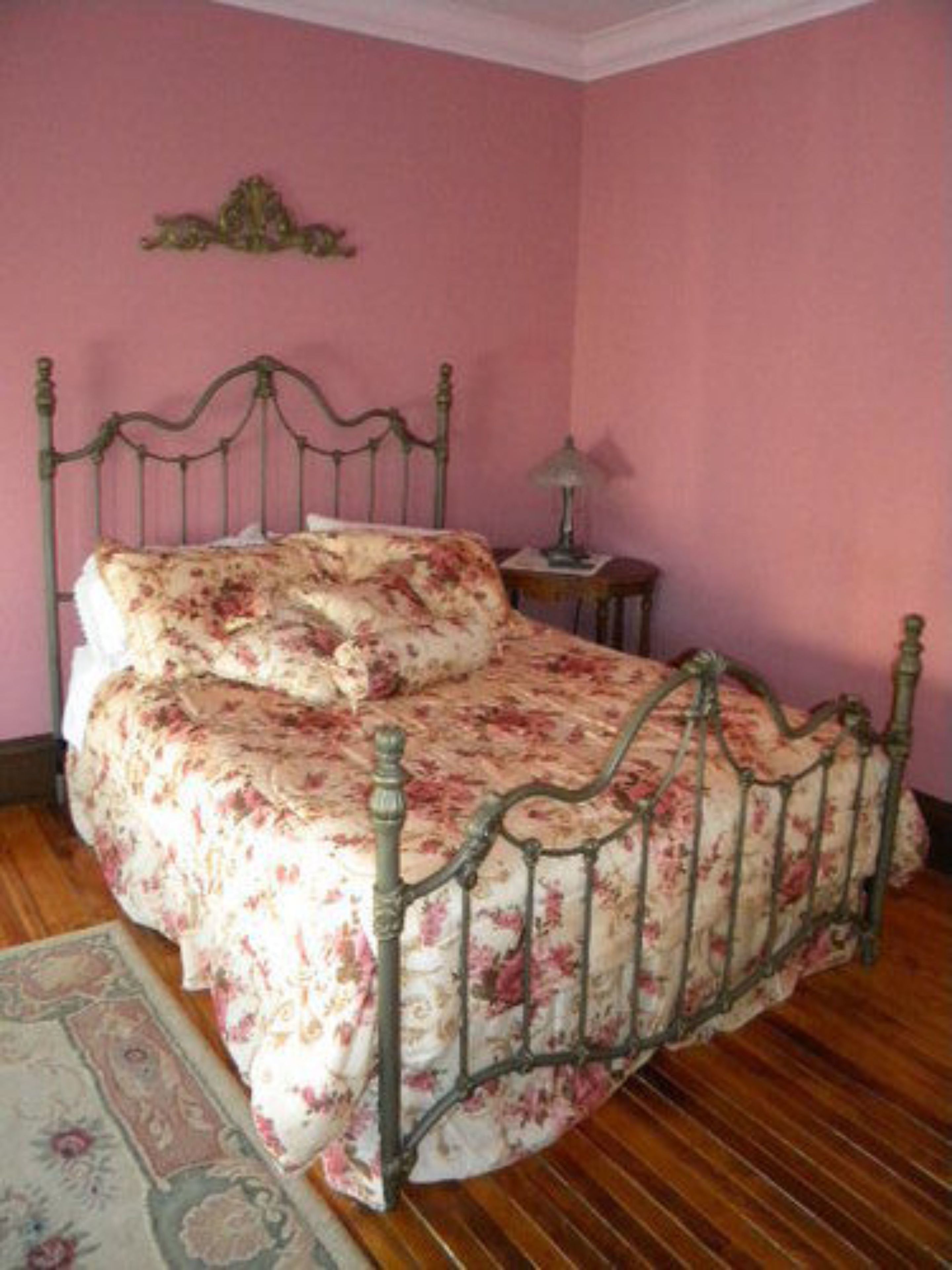 Un dormitorio con colcha estampada, estructura de cama metálica, mesita de noche pequeña con lámpara y un adorno en la pared.