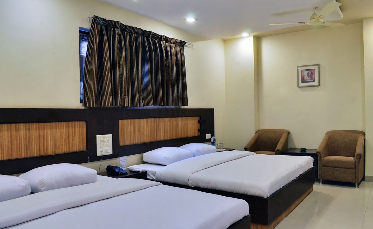 Quarto de hotel com duas camas de solteiro, cabeceira de madeira, ventilador de teto, uma mesinha, uma cadeira e janela com cortinas.