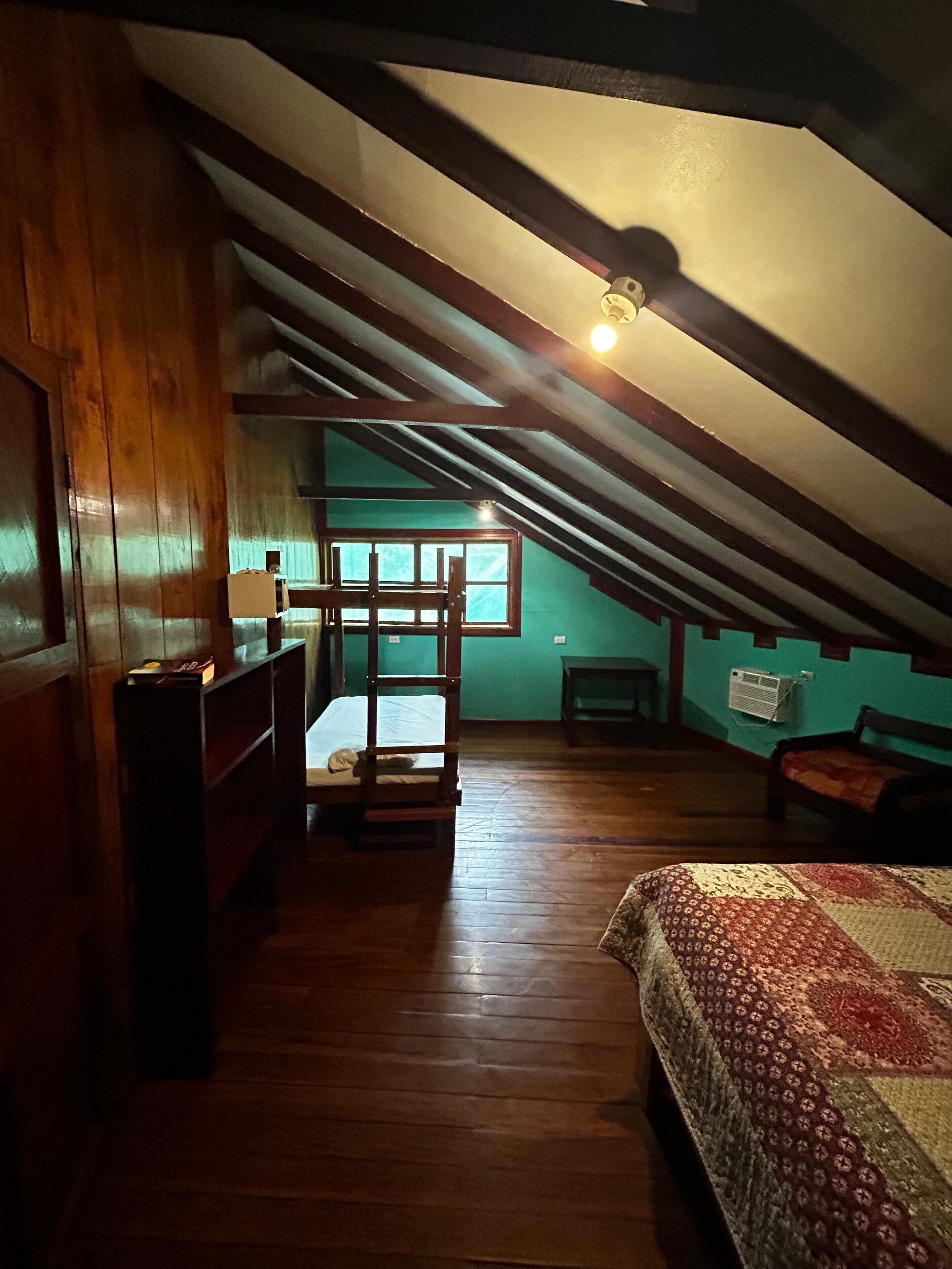 Une pièce avec un plafond en pente, des murs en bois, un lit recouvert d’une courtepointe à motifs, une petite table et une fenêtre donnant sur un paysage agréable.