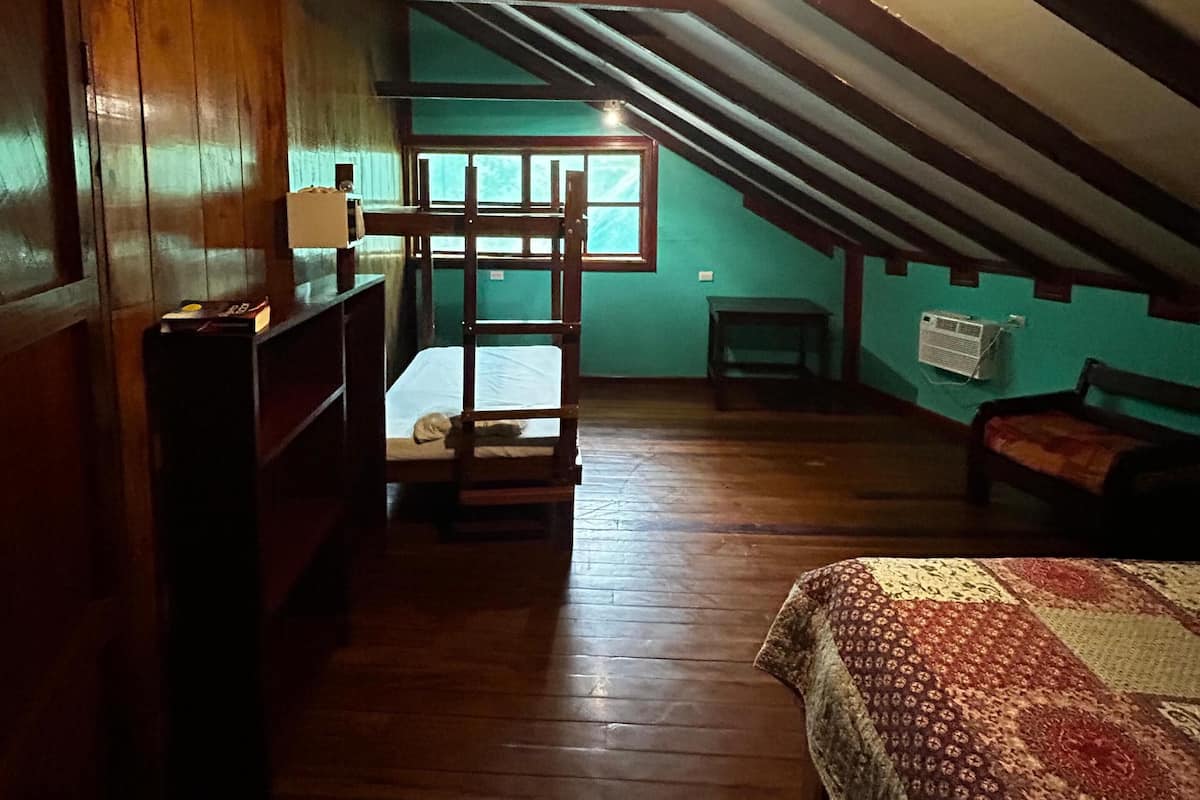 A room with a sloped ceiling, wooden walls, a bed with a patterned bedspread, a small table, and a window with a view.