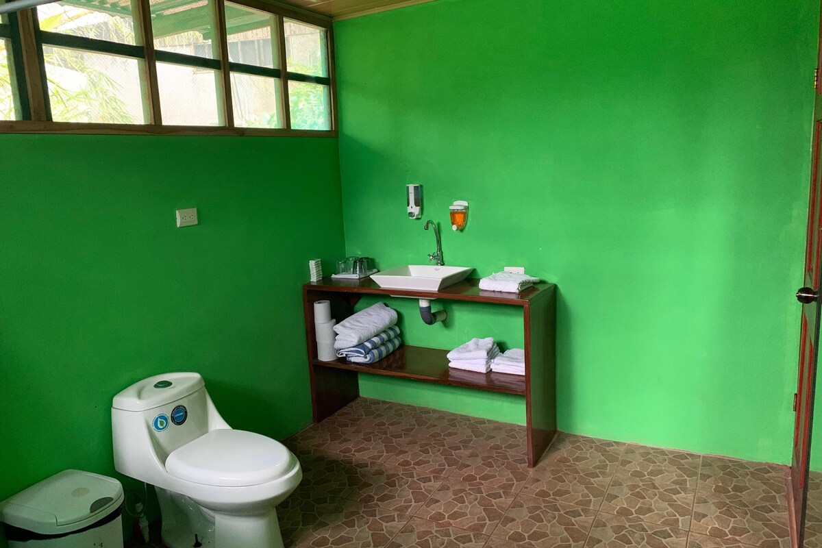 A bathroom with green walls, a white toilet, a sink with a mirror, and a shelf with towels.