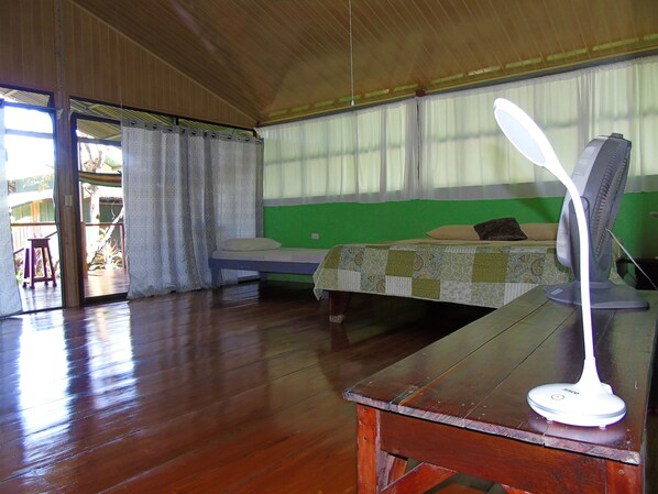 A room with a bed, a table with a fan, and a window with curtains.
