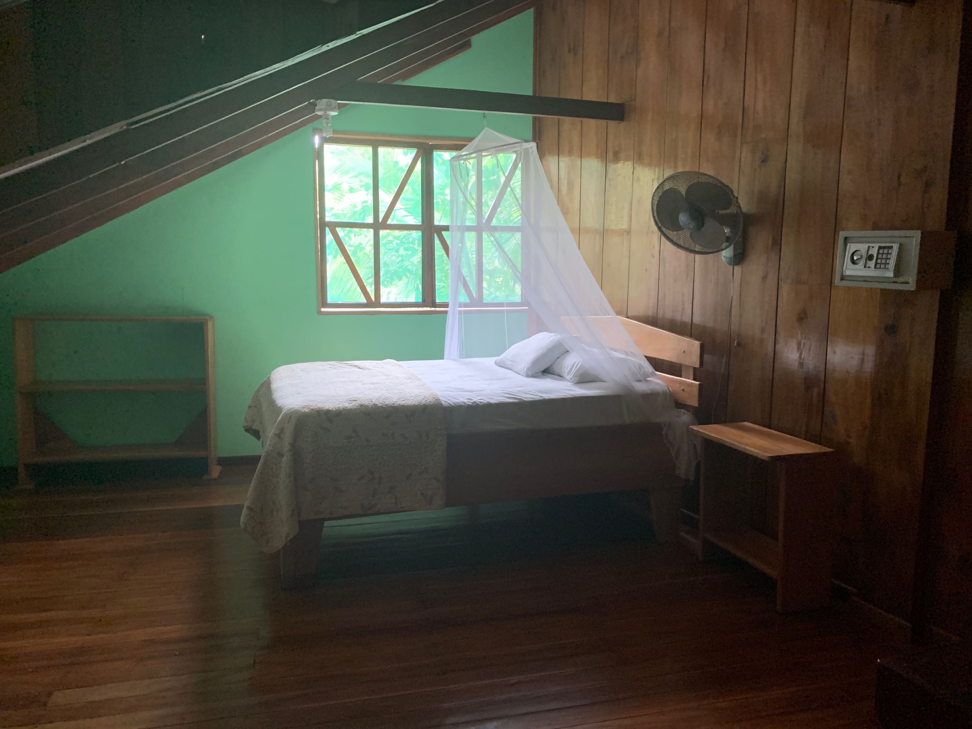Ein Zimmer mit einem Bett, einem Fenster mit weißem Vorhang, einer Holzbank und einem Wandventilator.