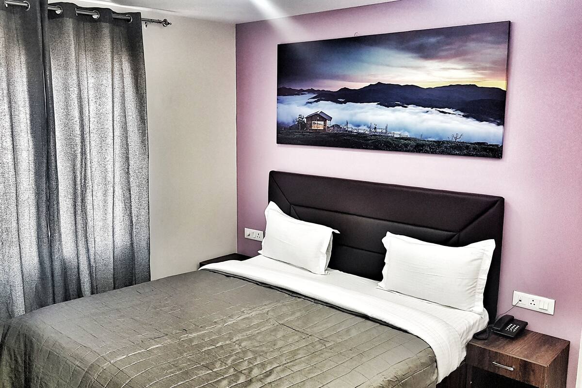 A bedroom with a bed, a nightstand, a painting of a mountain landscape, and a curtain.