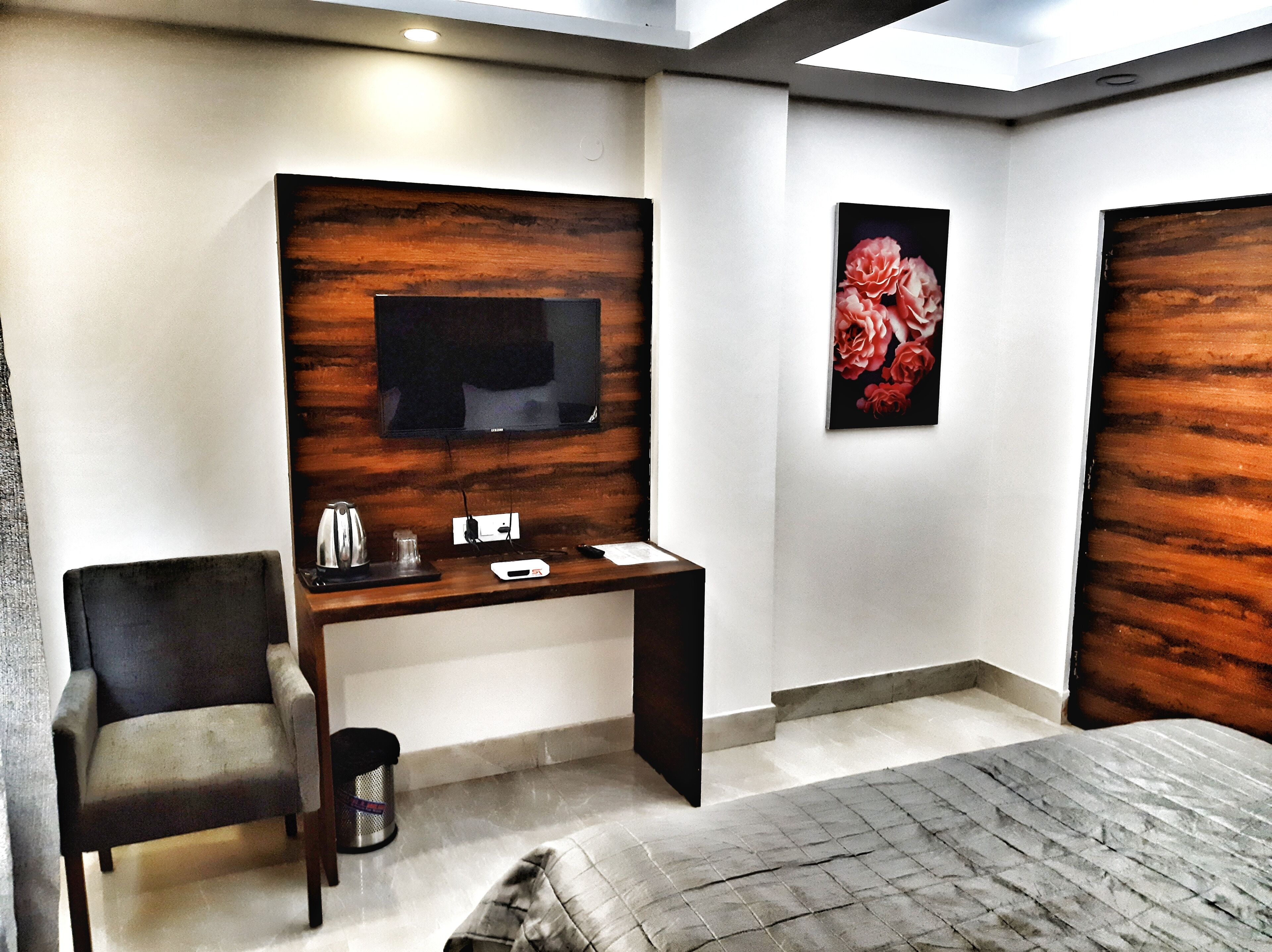 A modern hotel room with a wooden accent wall, a flat-screen TV mounted above a desk, a bed with a grey quilt, and a framed floral artwork on the wall.