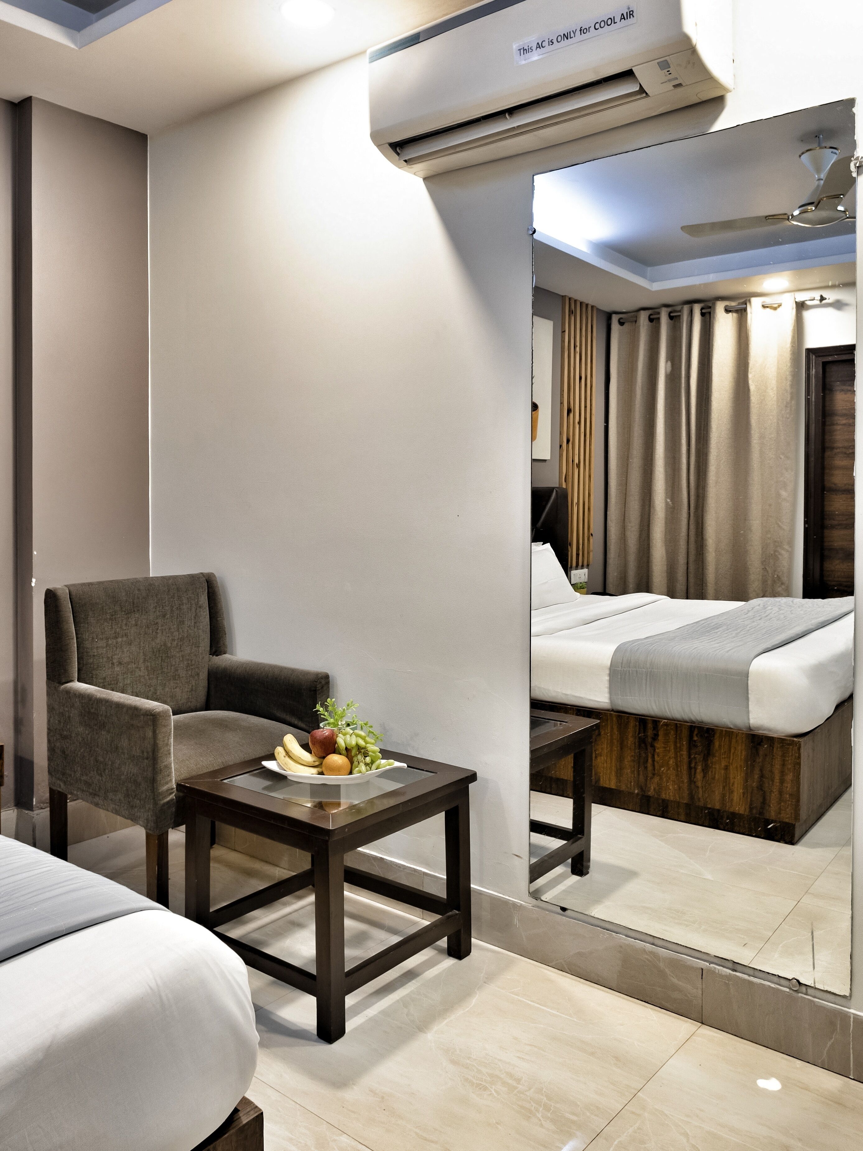 Une chambre d’hôtel moderne dotée d’un grand lit, d’un coin salon avec un fauteuil et une petite table, ainsi que d’un miroir qui reflète la pièce.