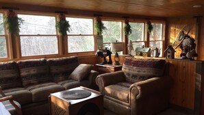 A cozy living room with a sofa, armchair, and a coffee table. There are windows with curtains and a wooden ceiling.