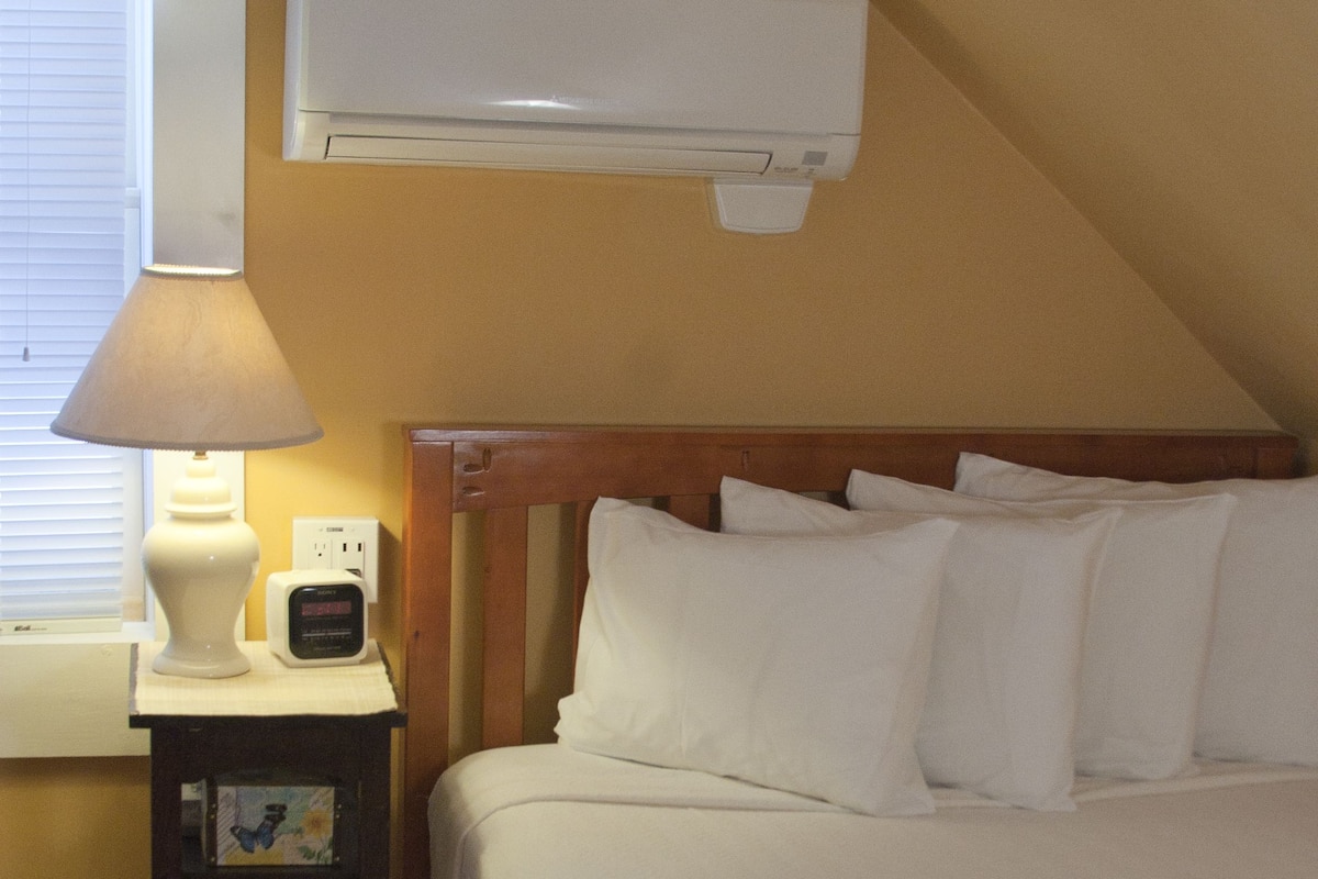 Quarto com cama, mesa de cabeceira com abajur, ar-condicionado na parede e janela com persianas.