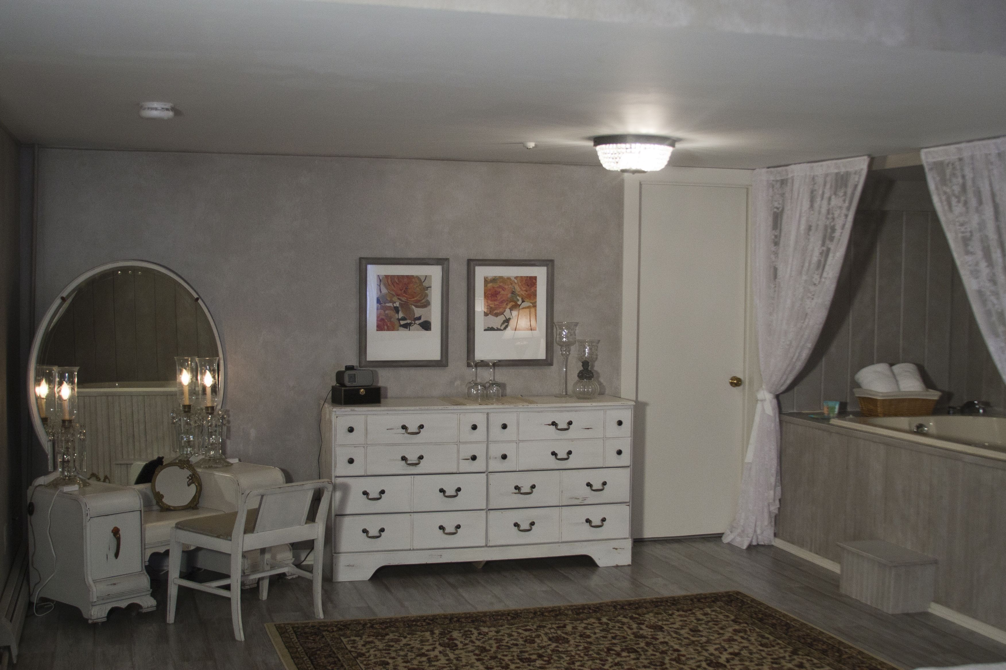 A bedroom with a vanity, dresser, and a large mirror. There are framed pictures on the wall, a chandelier, and a window with curtains.