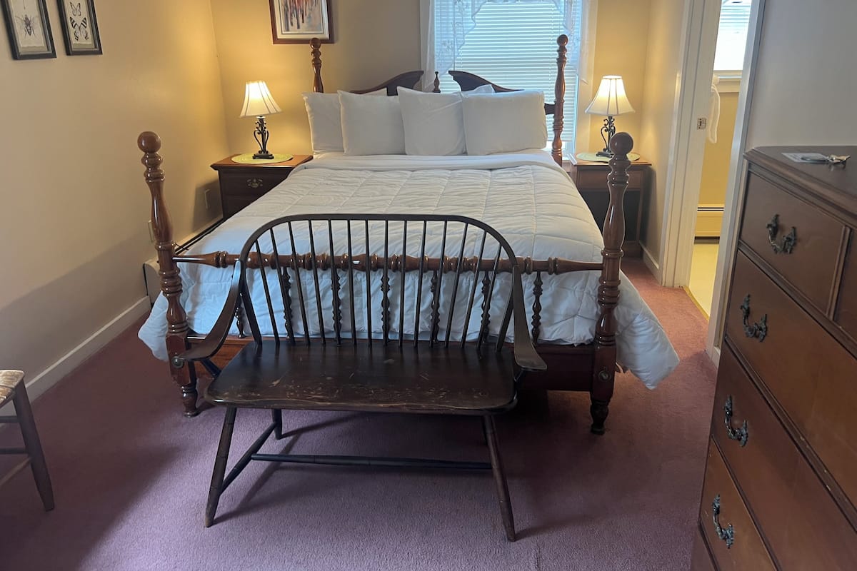 Quarto com cama de madeira com dossel, um banco, duas mesas de cabeceira com luminárias, cômoda e obras de arte emolduradas nas paredes.